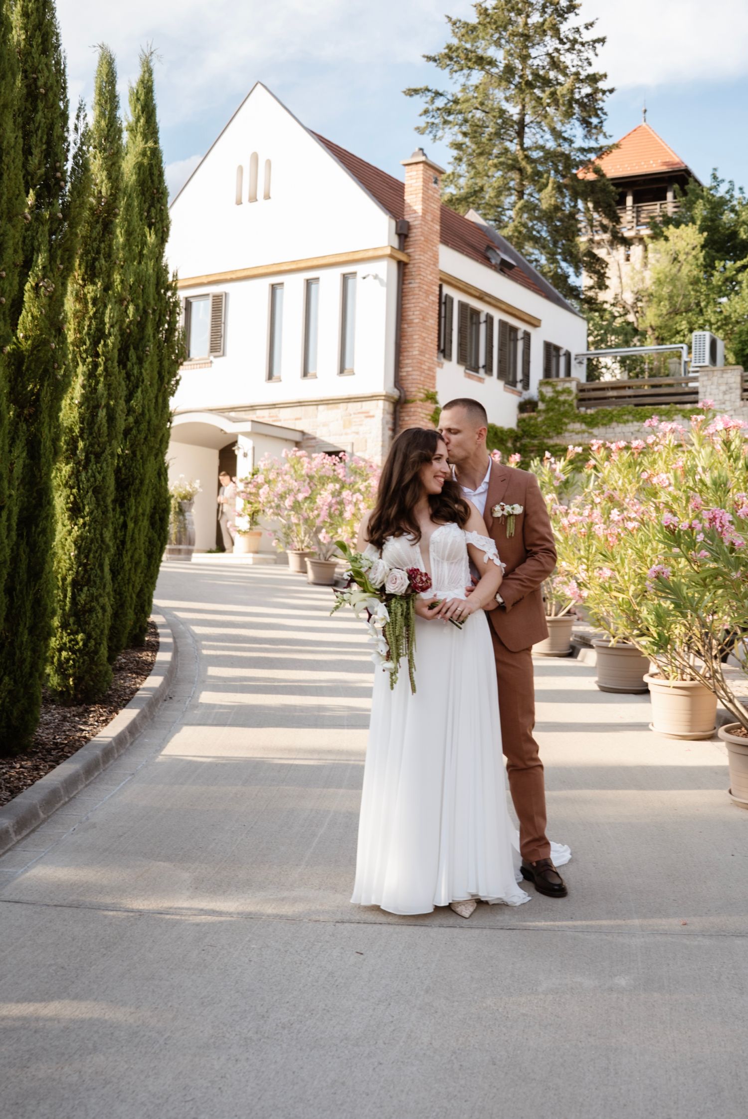 Bride and groom pose romantically in front of elegant white villa surrounded by tall cypress trees.