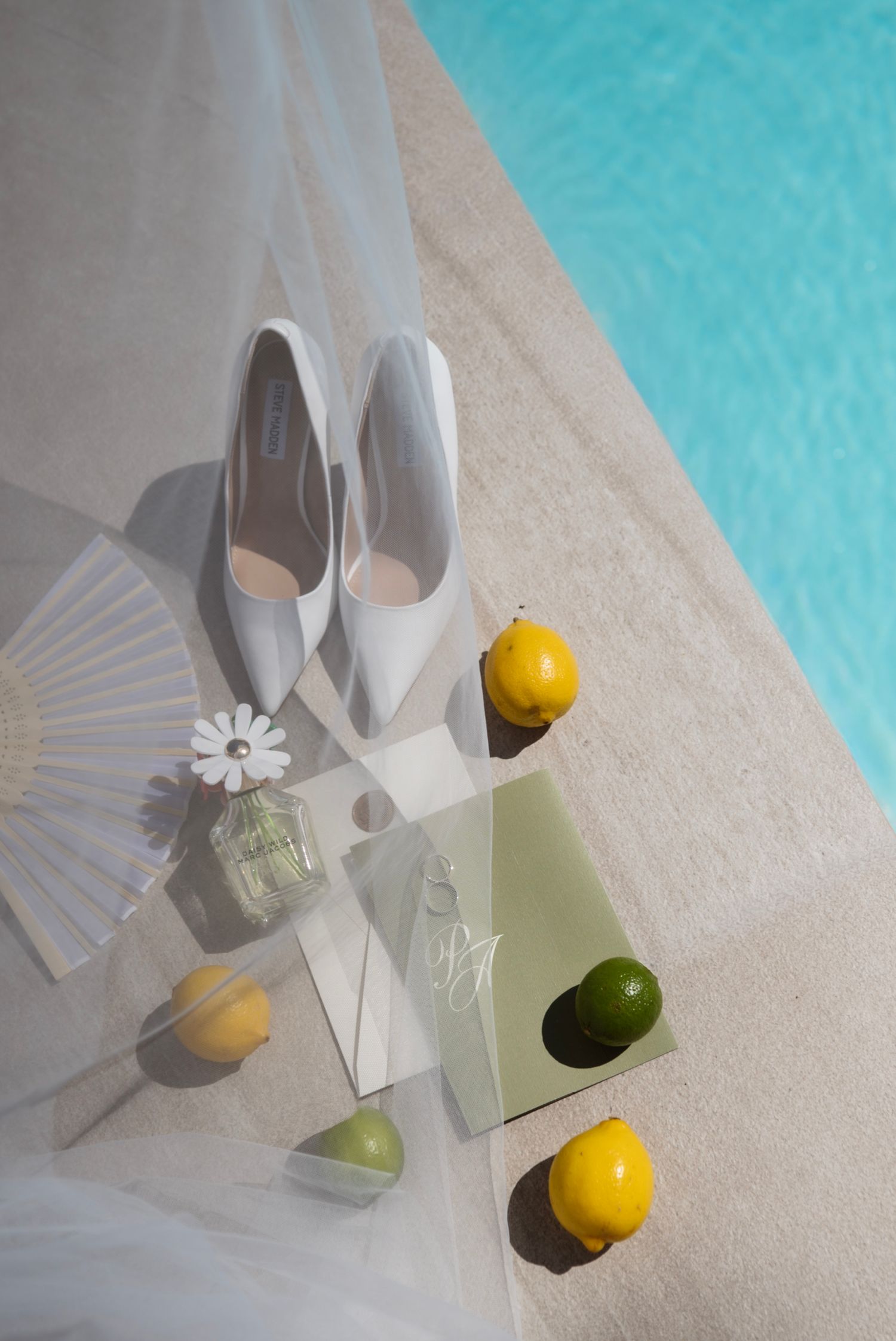 White bridal heels beside lemons, lime, perfume, and a fan on a mirrored tray by a pool.