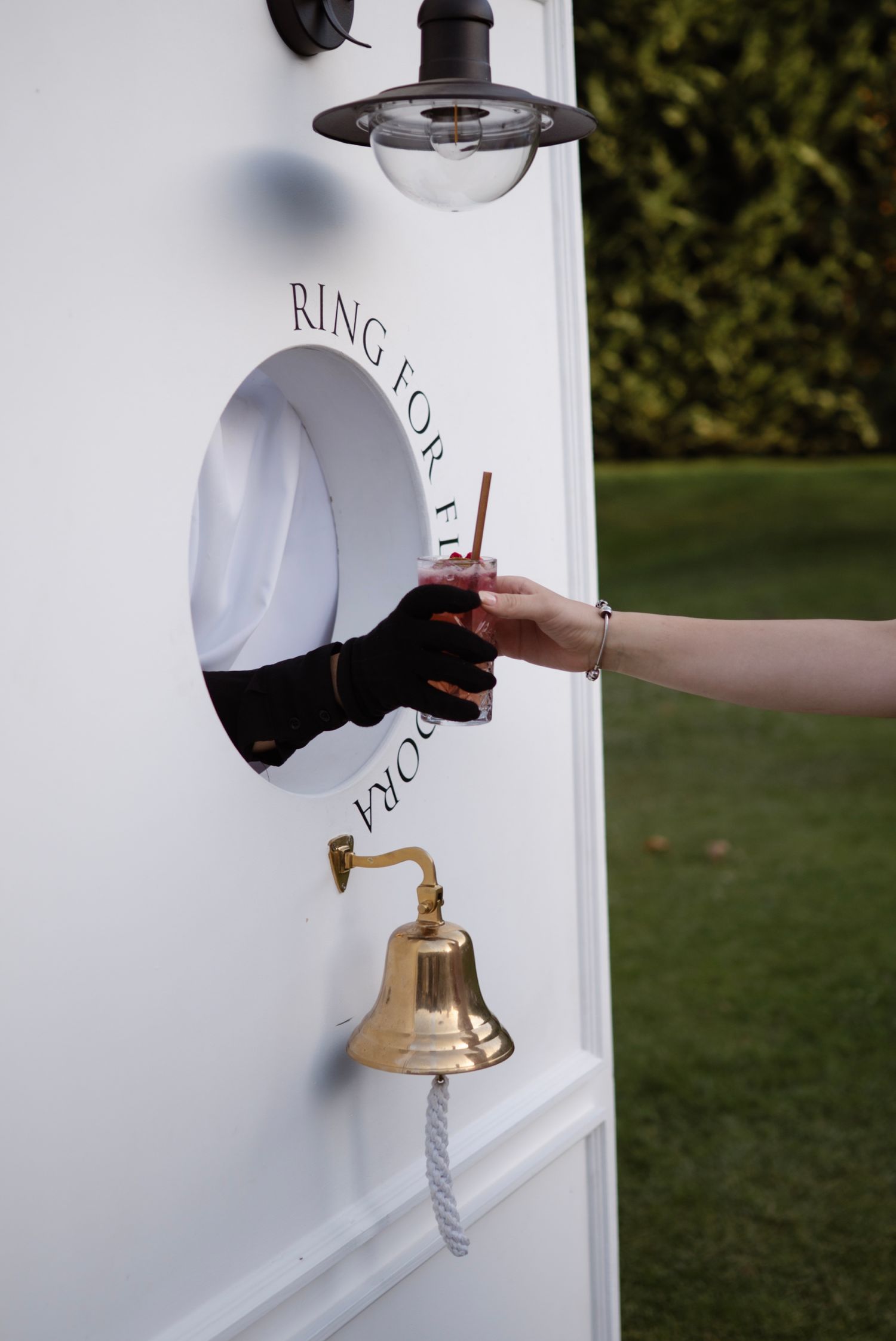 A brass bell and circular window on a white bar cart with 'Ring for Prosecco' text, with a hand receiving a drink.