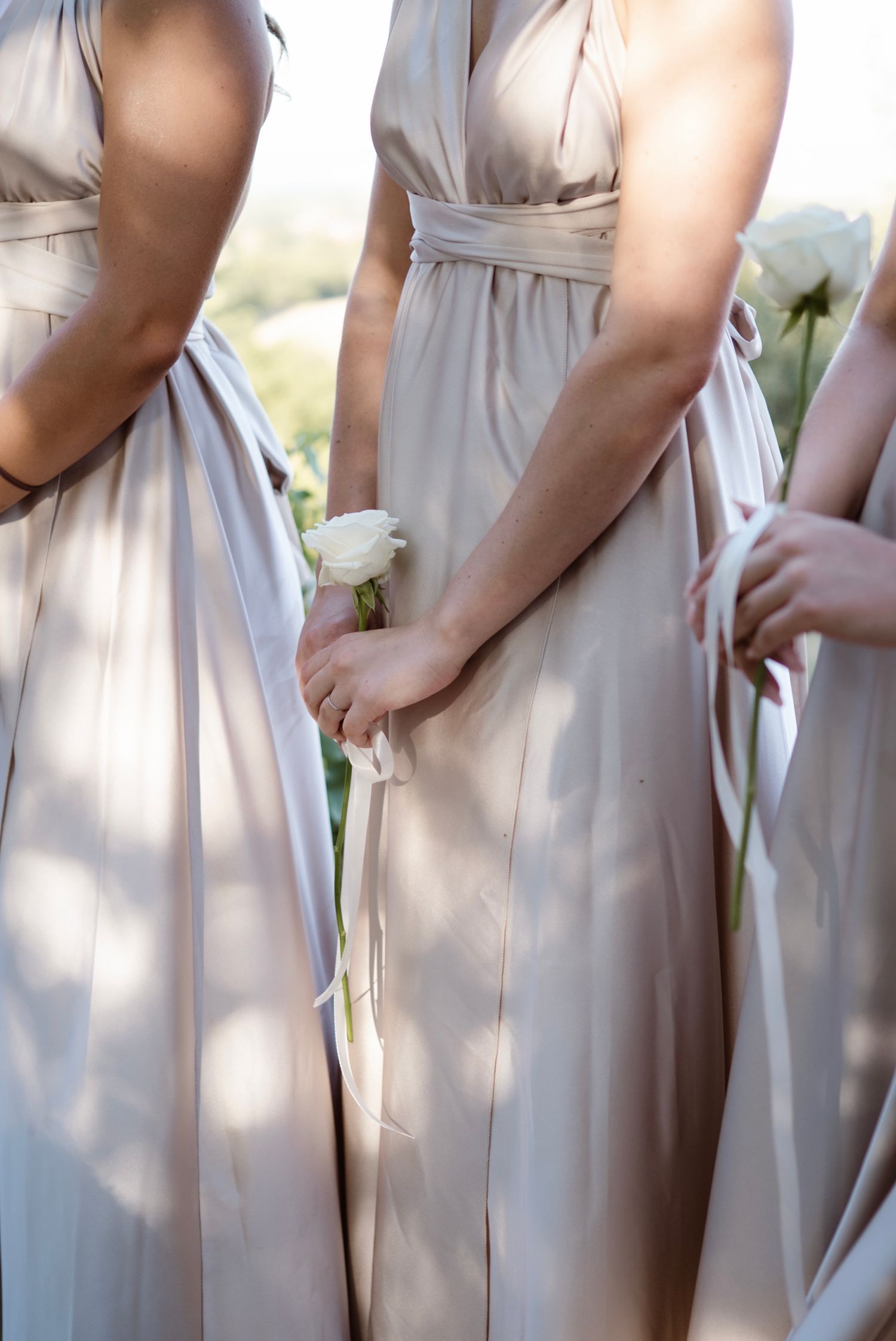 Bridesmaids in elegant champagne satin wrap gowns holding white flowers at an outdoor wedding.
