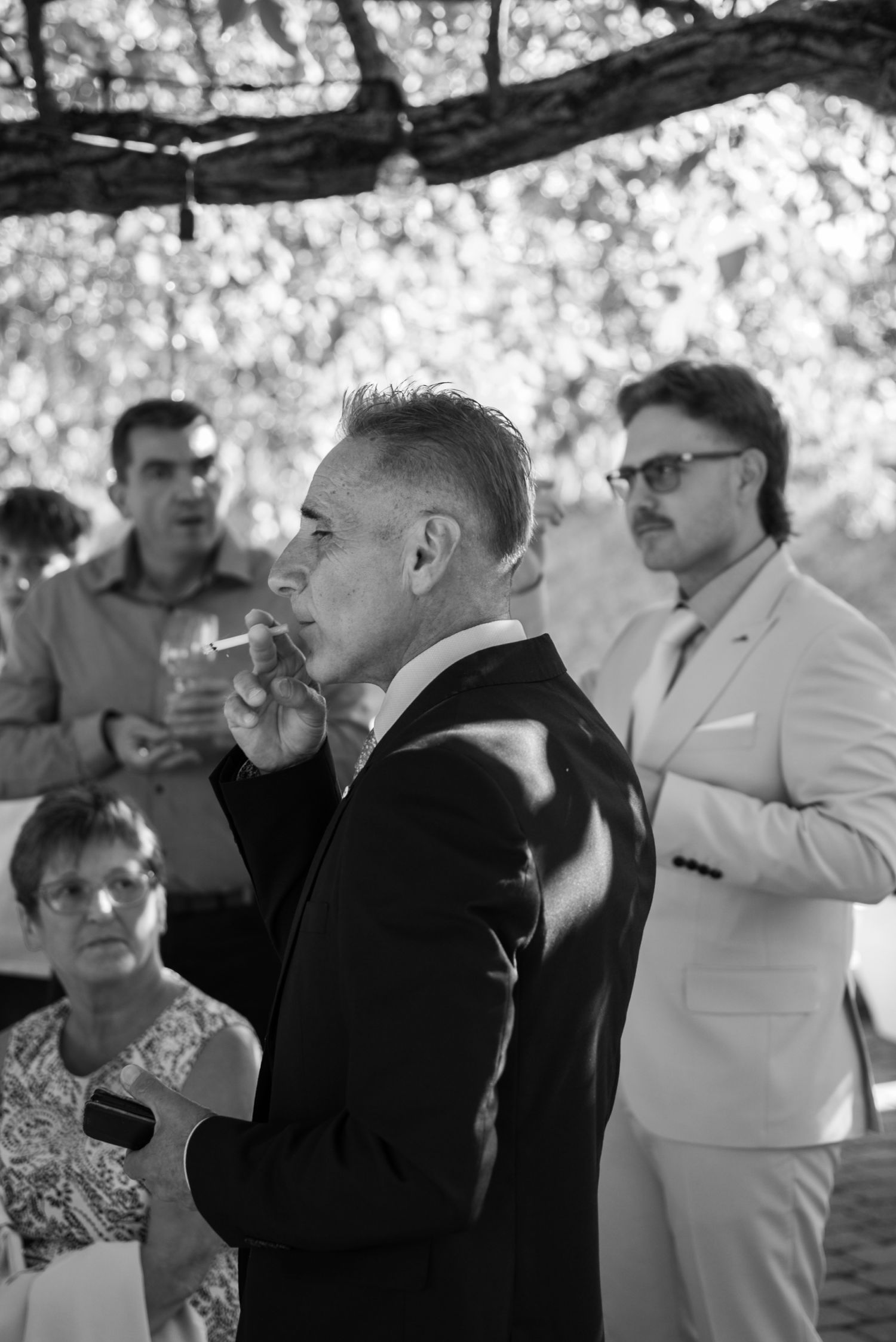 Black and white photo of a man in a suit smoking a cigar at an outdoor event with guests in the background.