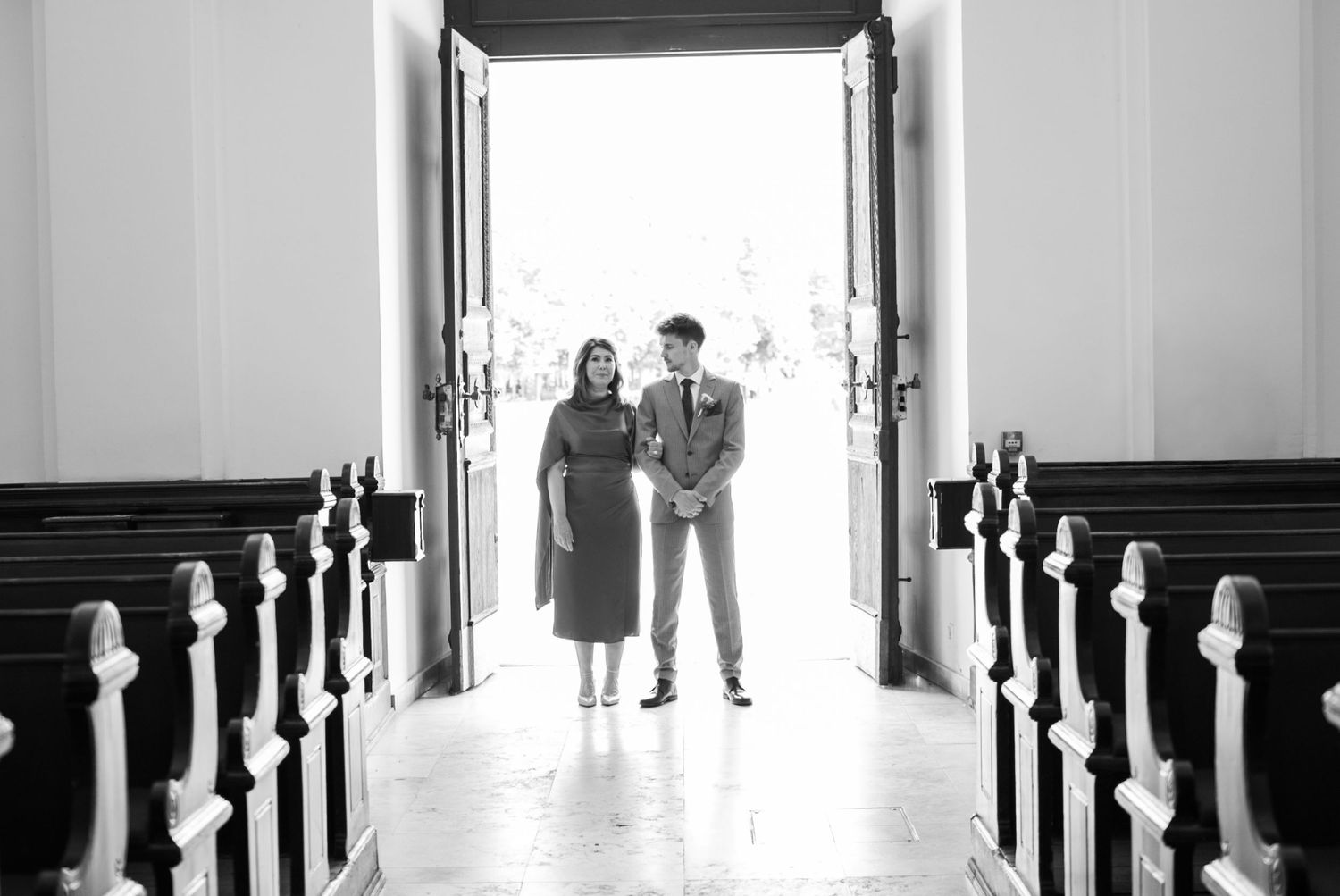 Black and white photo of a couple standing in church doorway, flanked by wooden pews, in elegant wedding attire.