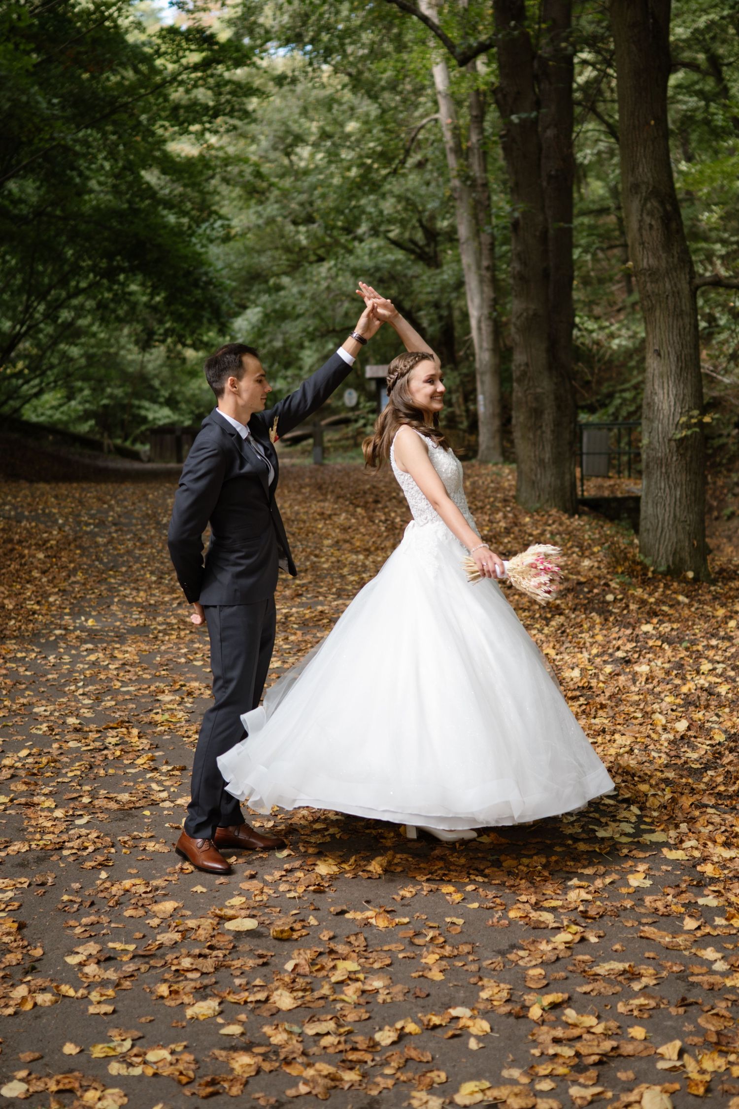 A couple dances playfully on a path covered in fallen autumn leaves.