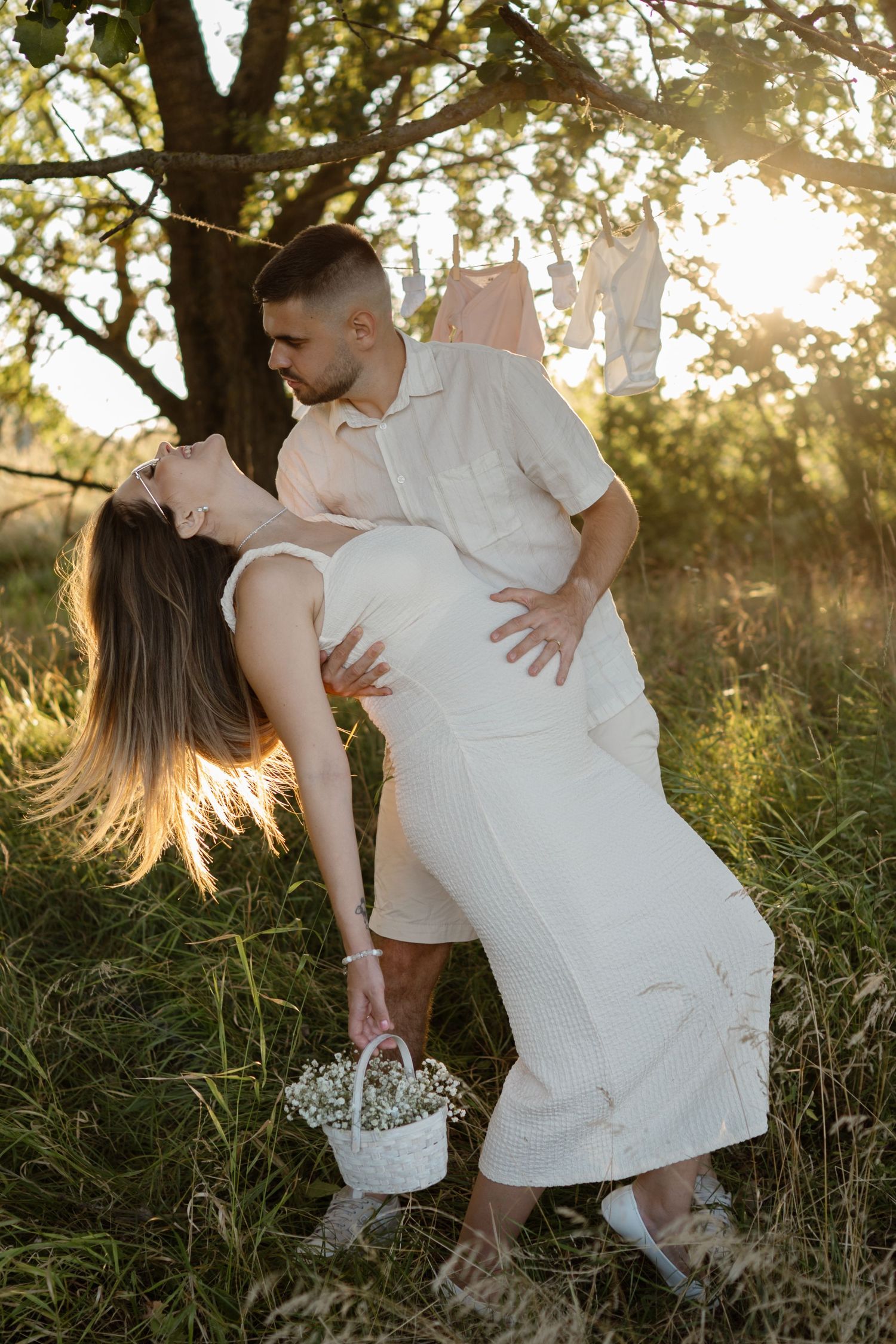 A romantic dip pose between a couple in white outfits under a tree at sunset.