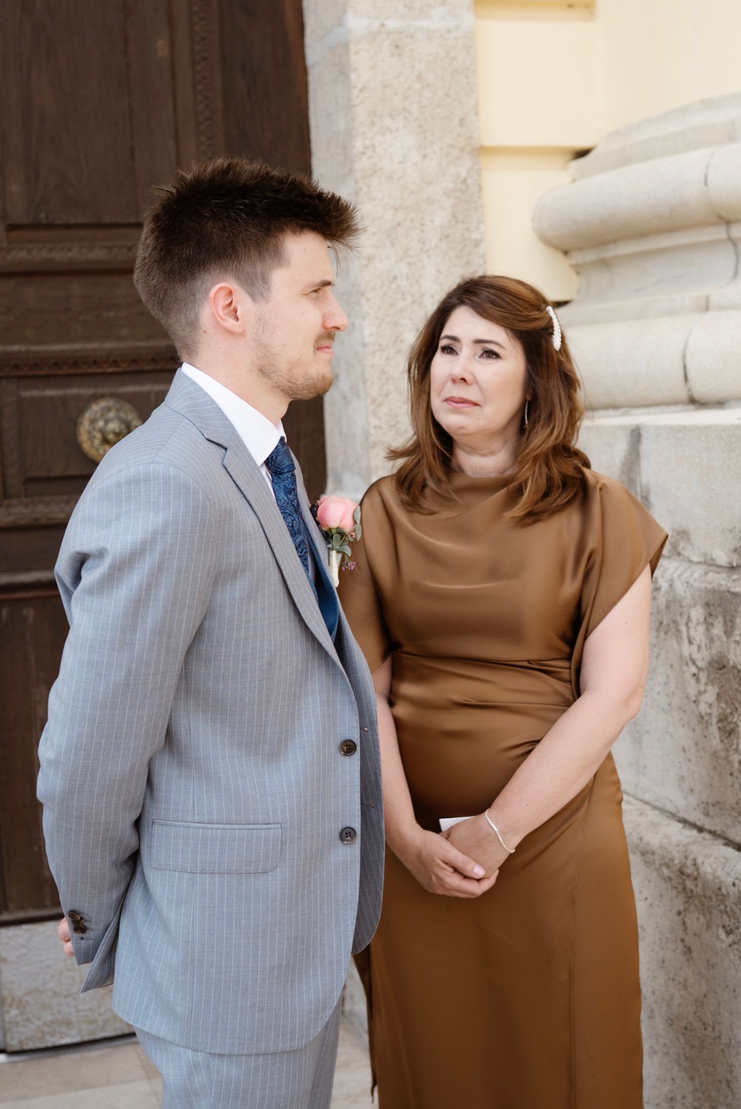 A man in a gray suit and a woman in a brown dress stand together near a stone column and wooden door.