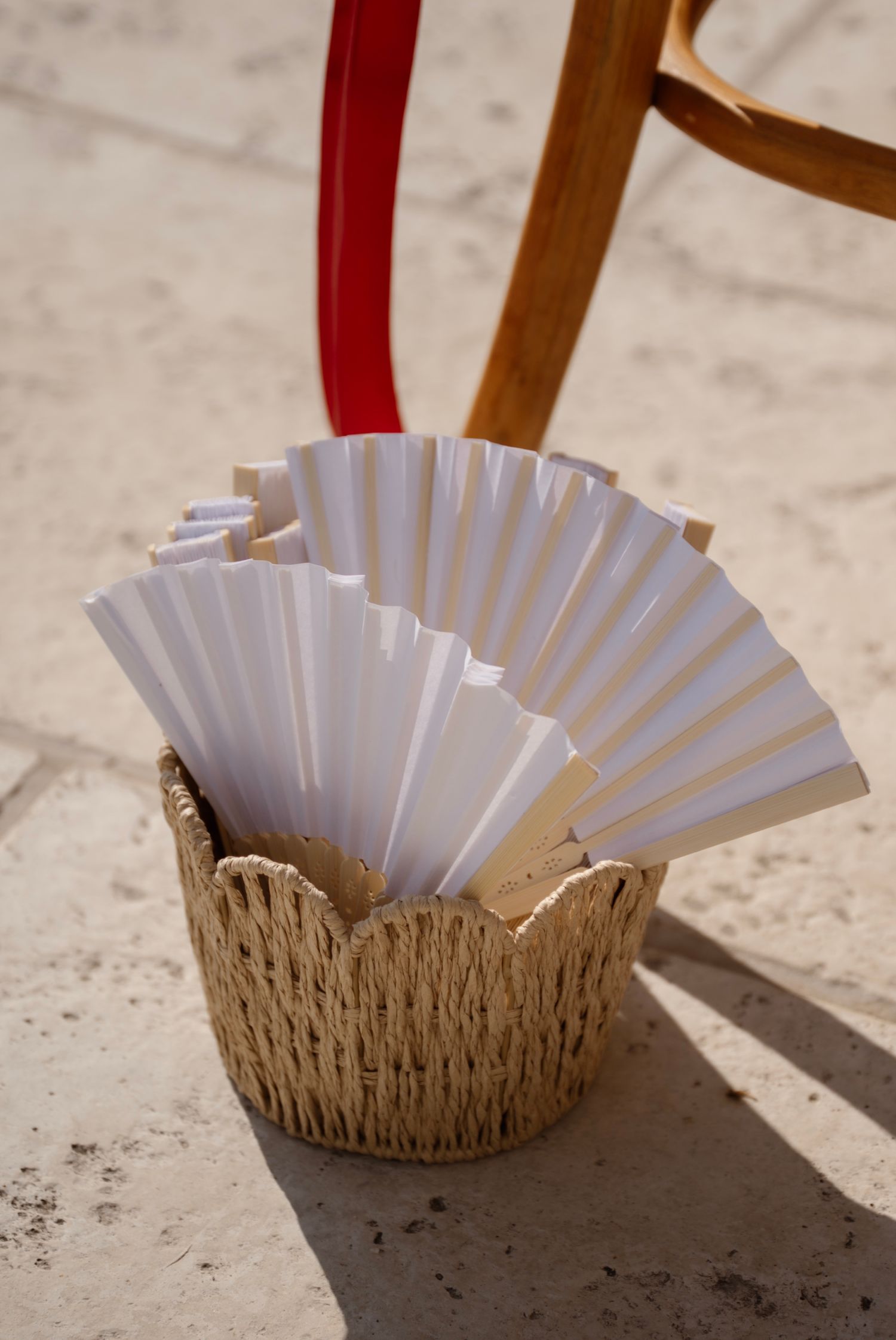 White paper hand fans displayed in a woven straw basket outdoors at a wedding ceremony.