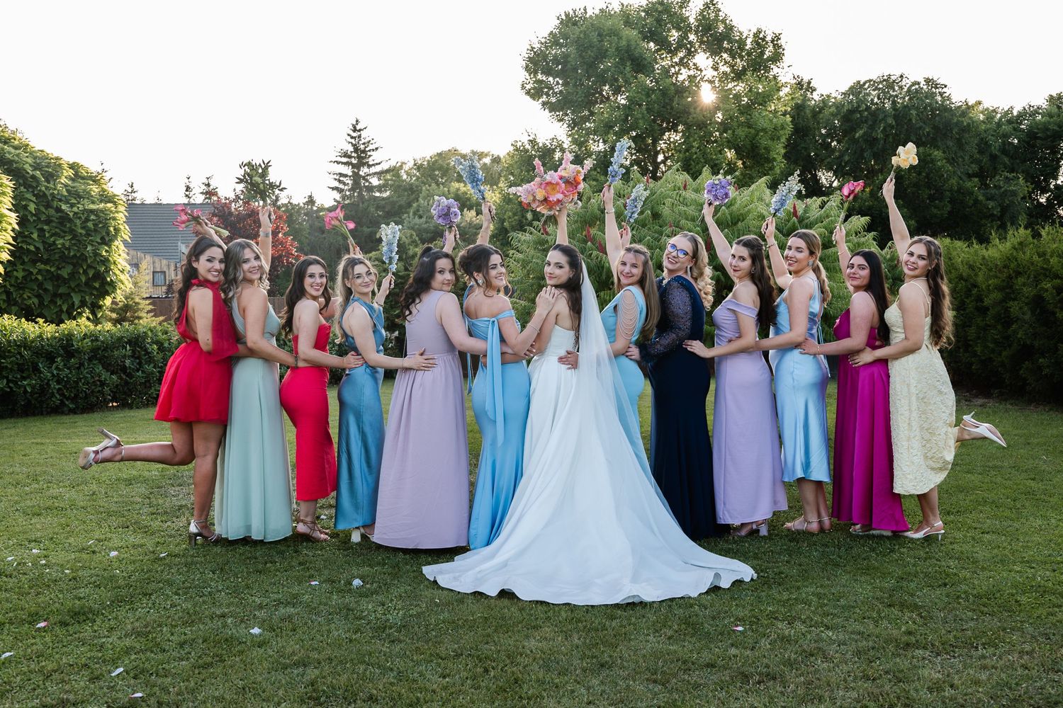 Wedding party in colorful formal dresses posed together on a green lawn during sunset.