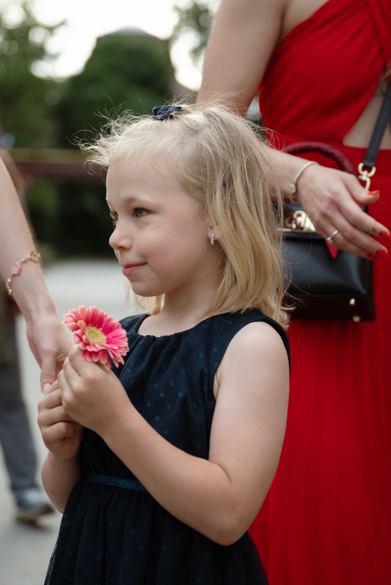 A young blonde girl in a navy dress holds a pink gerbera daisy at an outdoor wedding event.