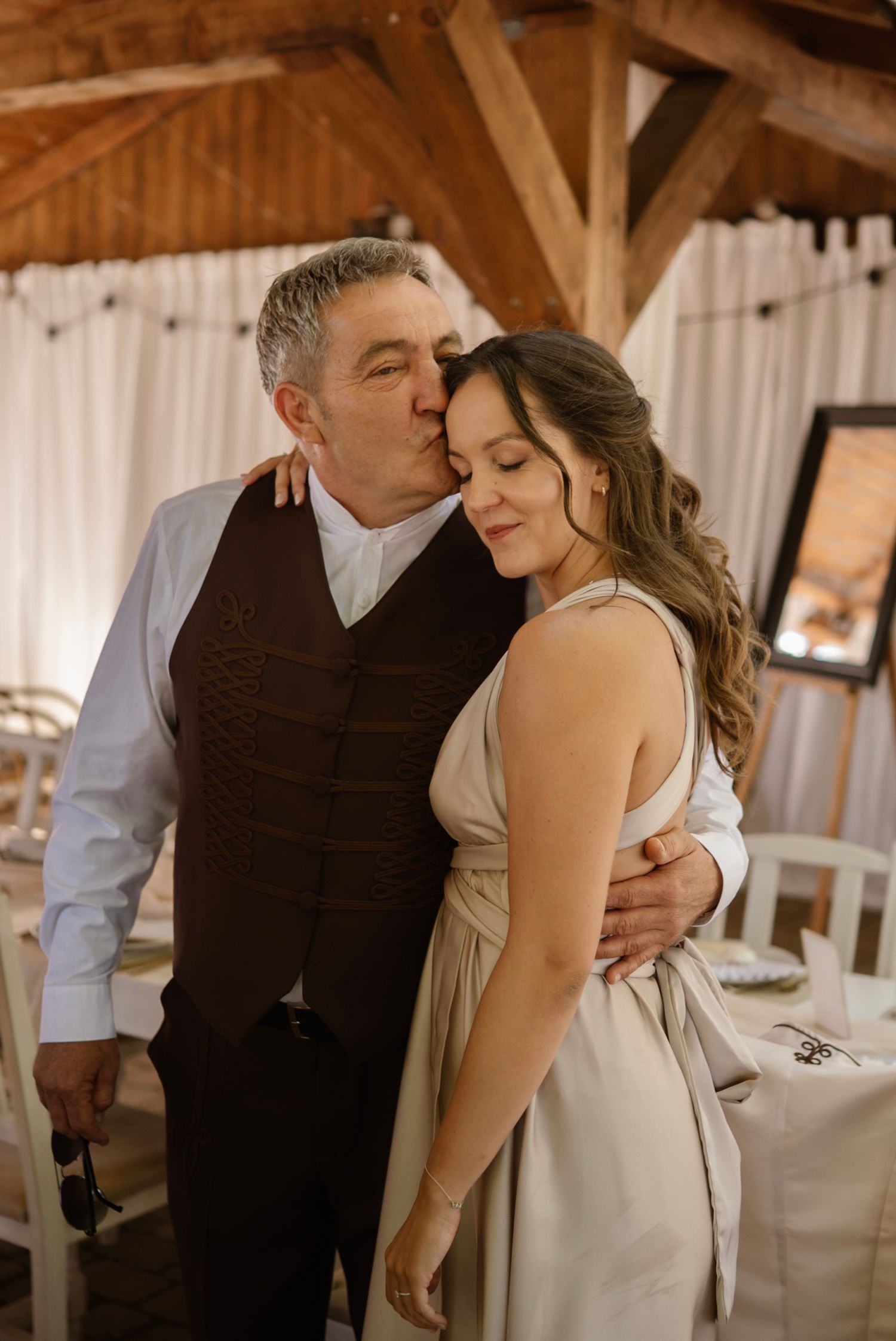 An older man in a brown vest kisses a young woman in a beige dress at an elegant wedding venue.