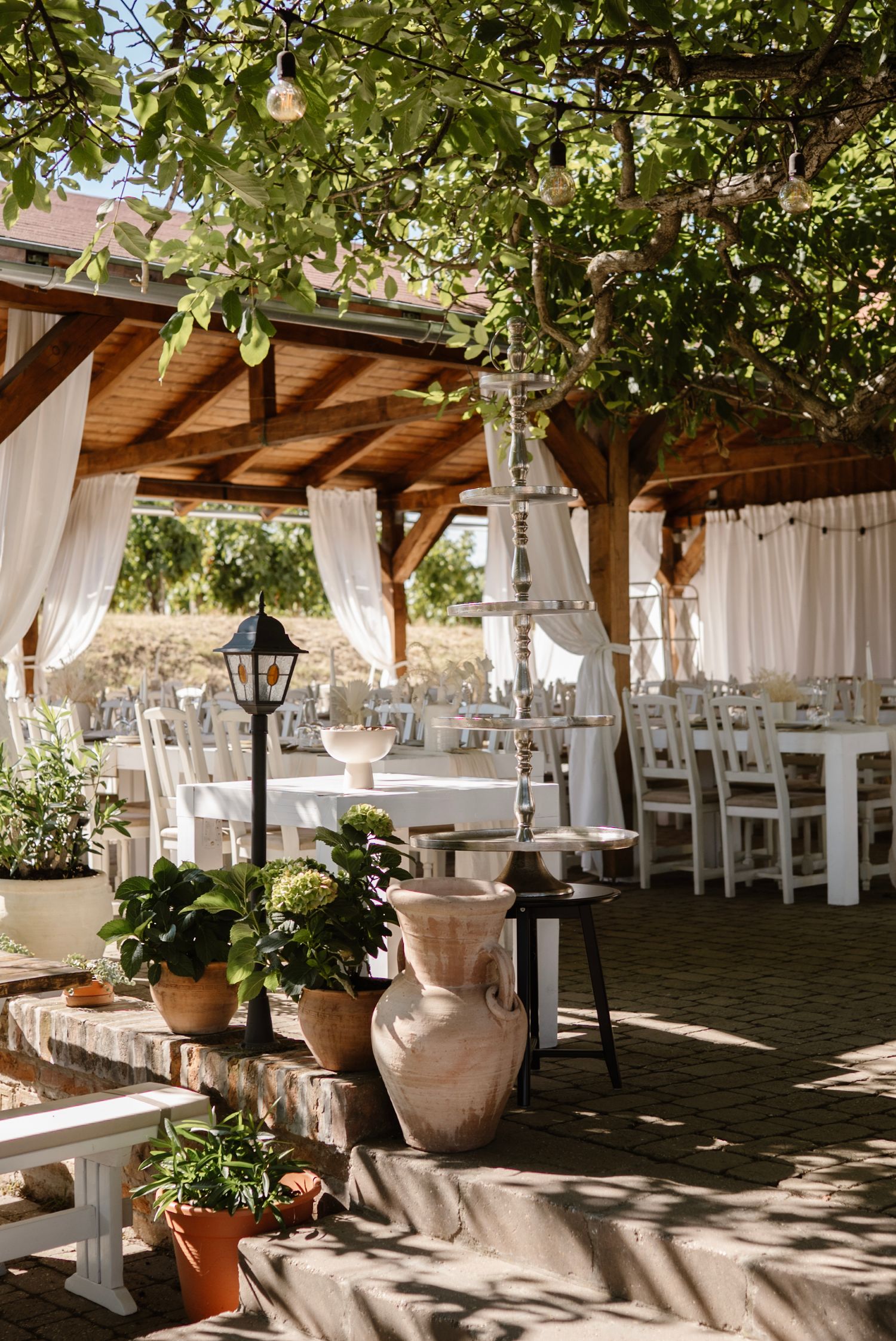 Elegant outdoor wedding reception under wooden pergola with white drapes, string lights, and terracotta pots.