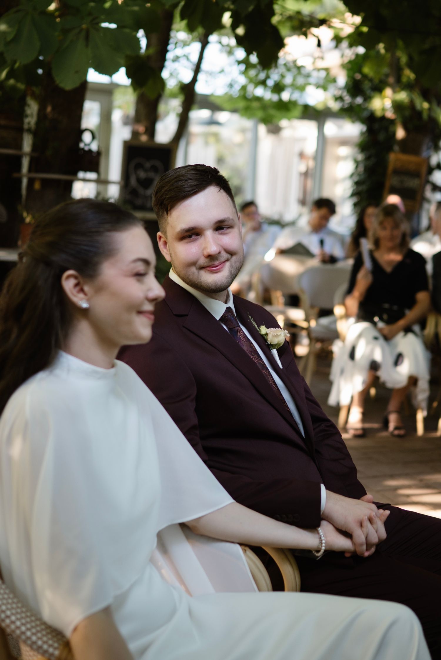 Smiling groom in burgundy suit holds bride's hand while seated at outdoor wedding ceremony under green trees.