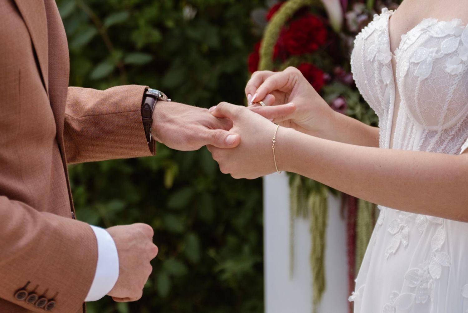 Groom placing a wedding ring on bride's finger during outdoor ceremony with floral arch backdrop.
