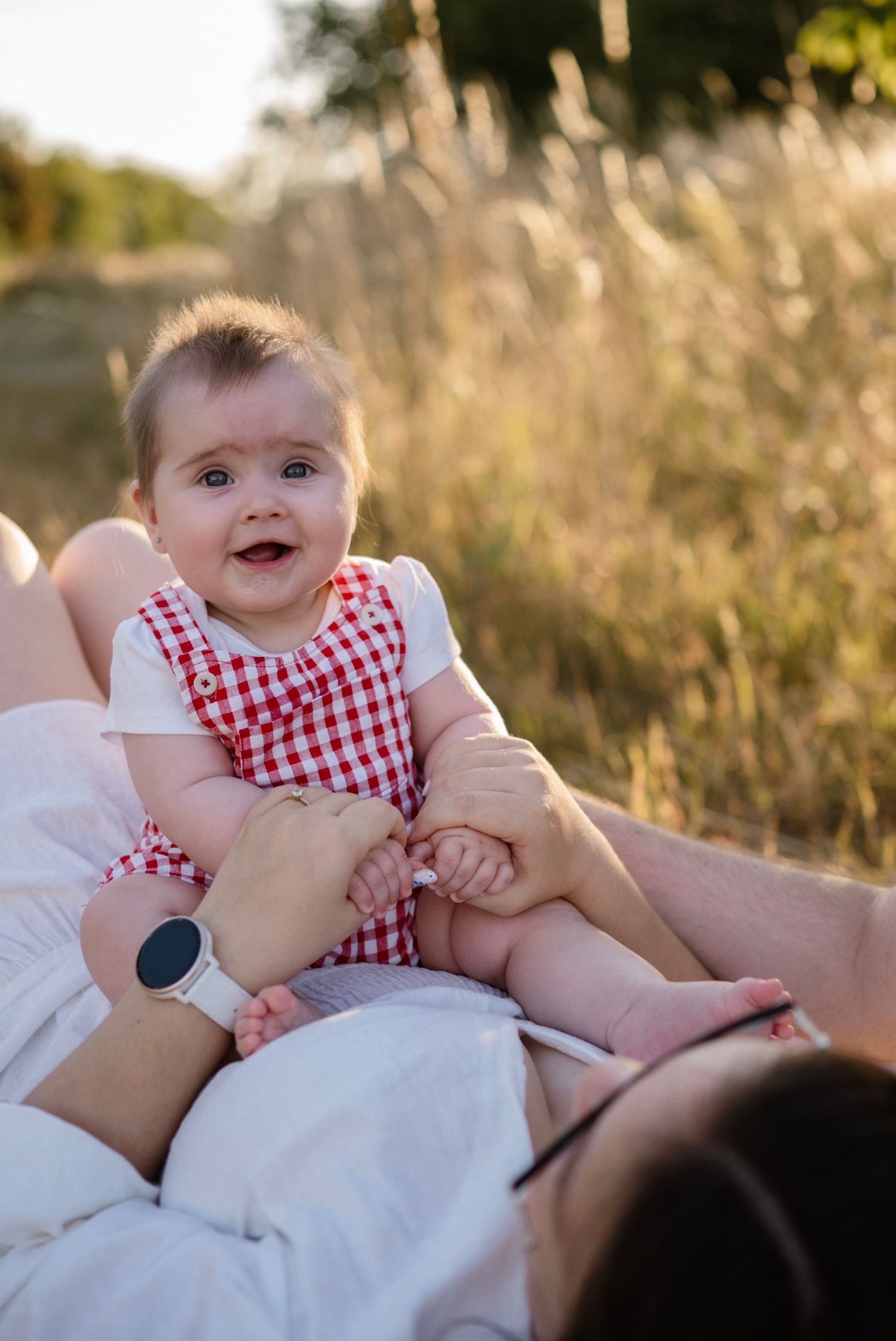 Adorable baby in red gingham outfit smiling outdoors, held by parent in golden field at sunset.