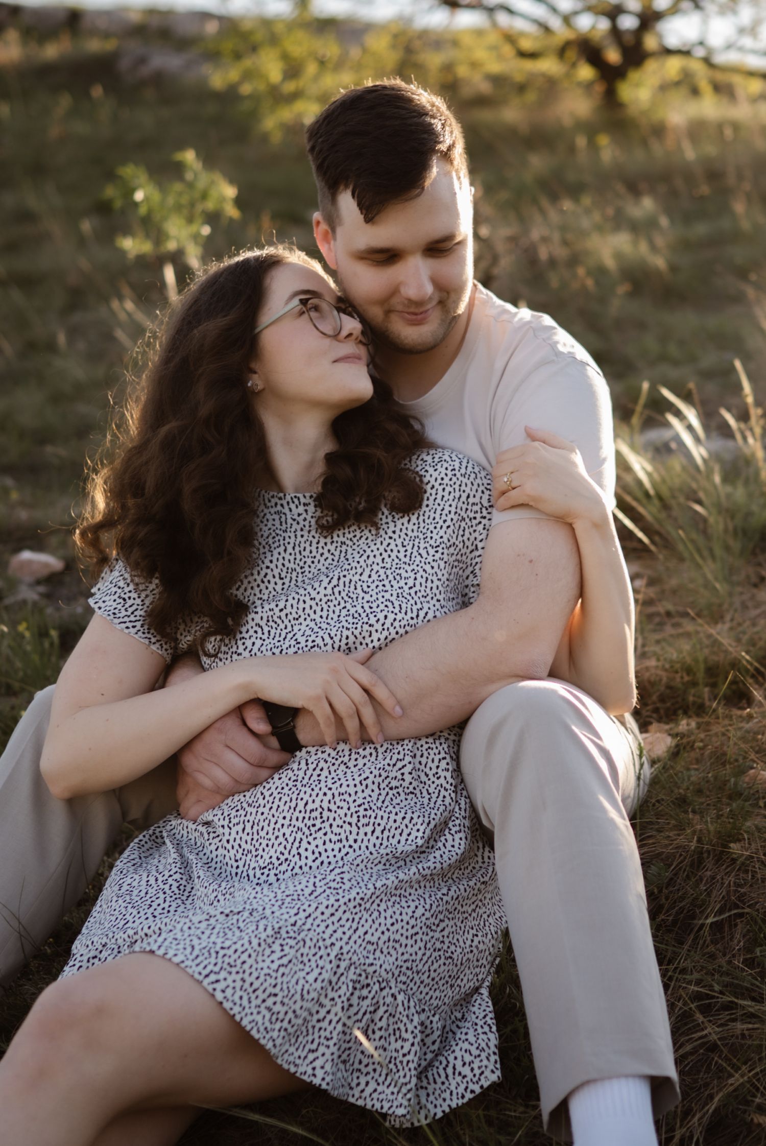 A couple sits together outdoors at golden hour, woman in floral dress leaning back into mans embrace smiling up at him.