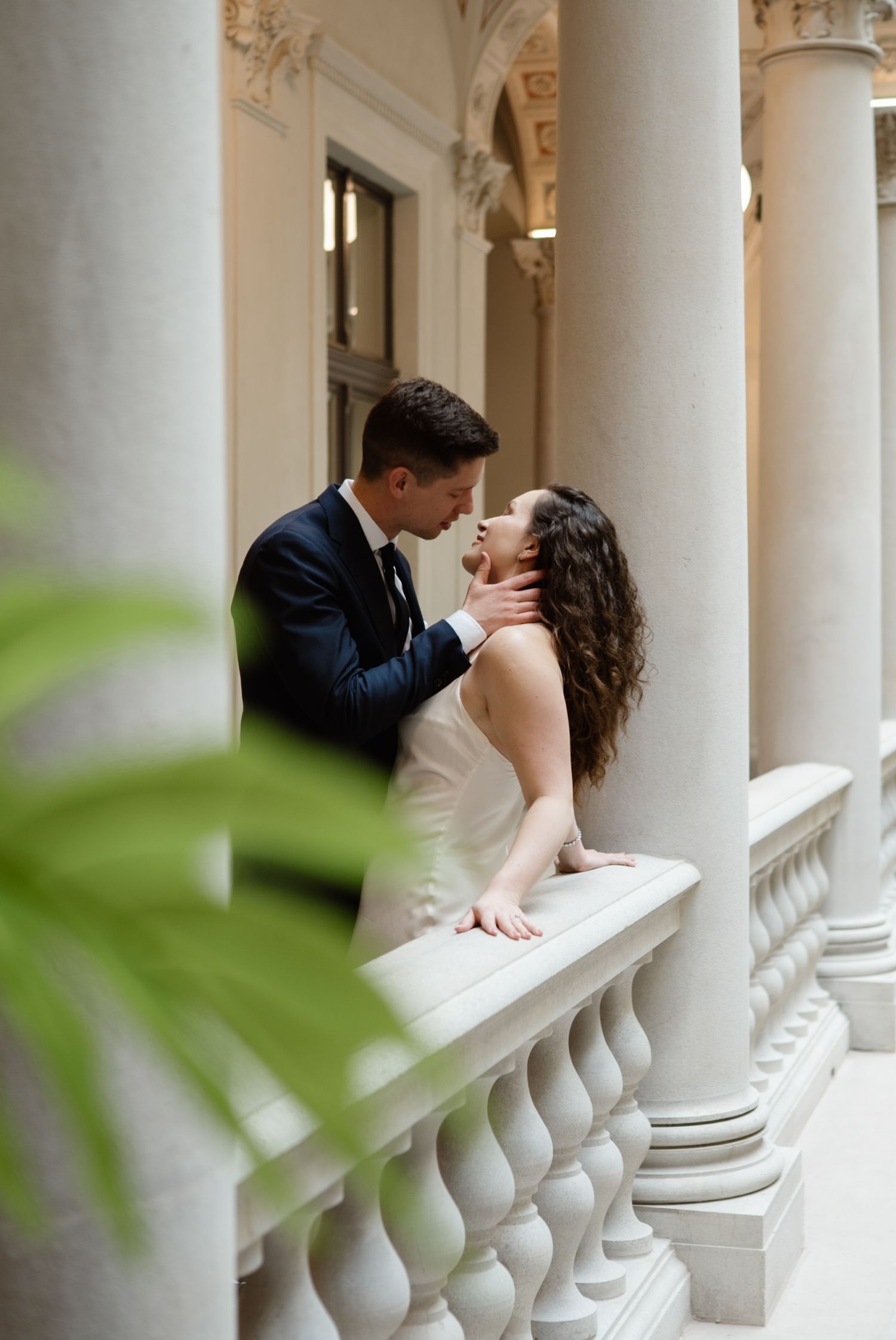 Bride and groom share a romantic moment among elegant white columns at a classical wedding venue.