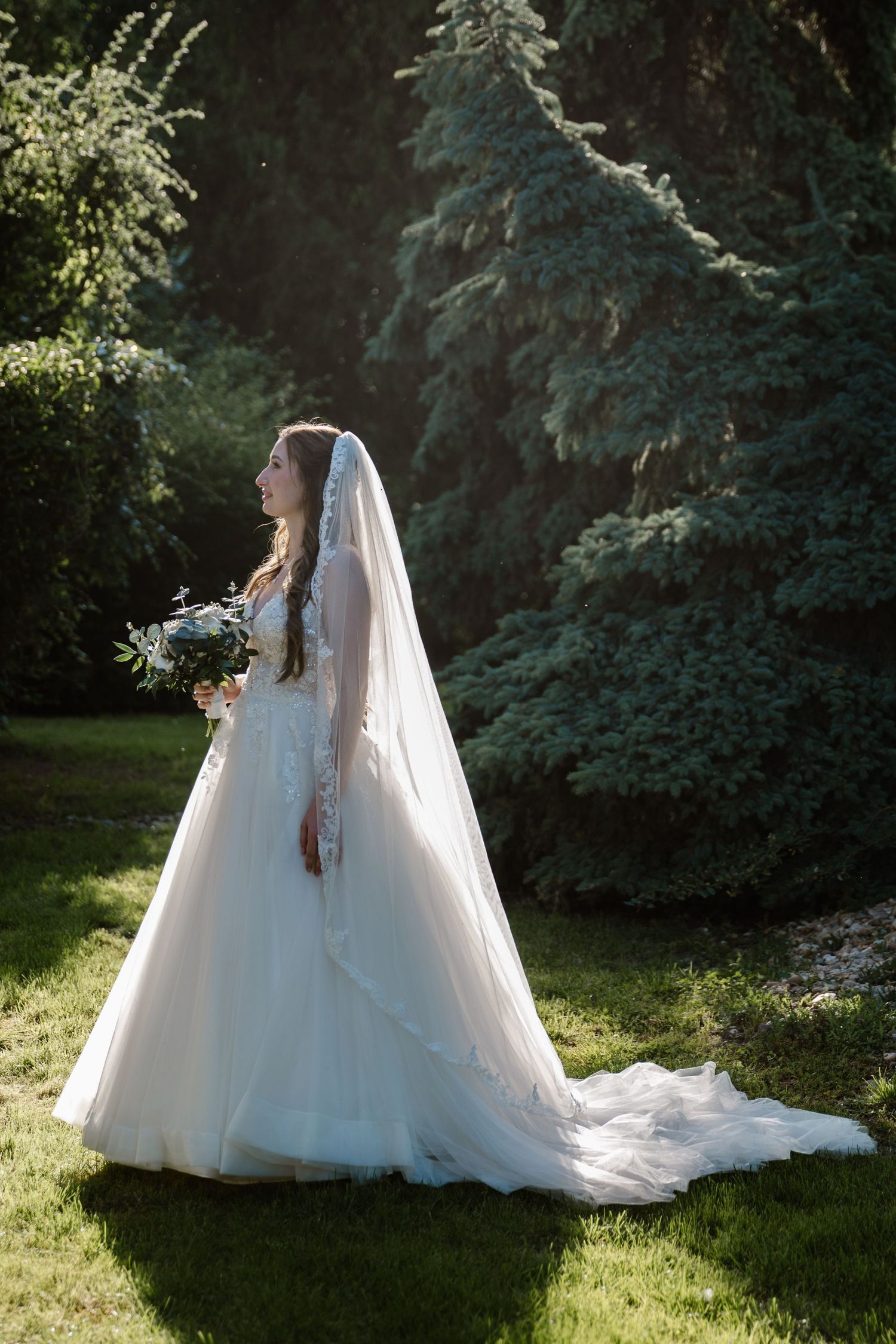 A bride in a flowing white gown poses in a garden with evergreen trees.
