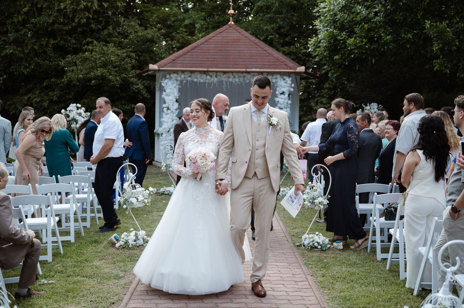 Outdoor wedding ceremony setup with white chairs and guests gathered on a lawn.