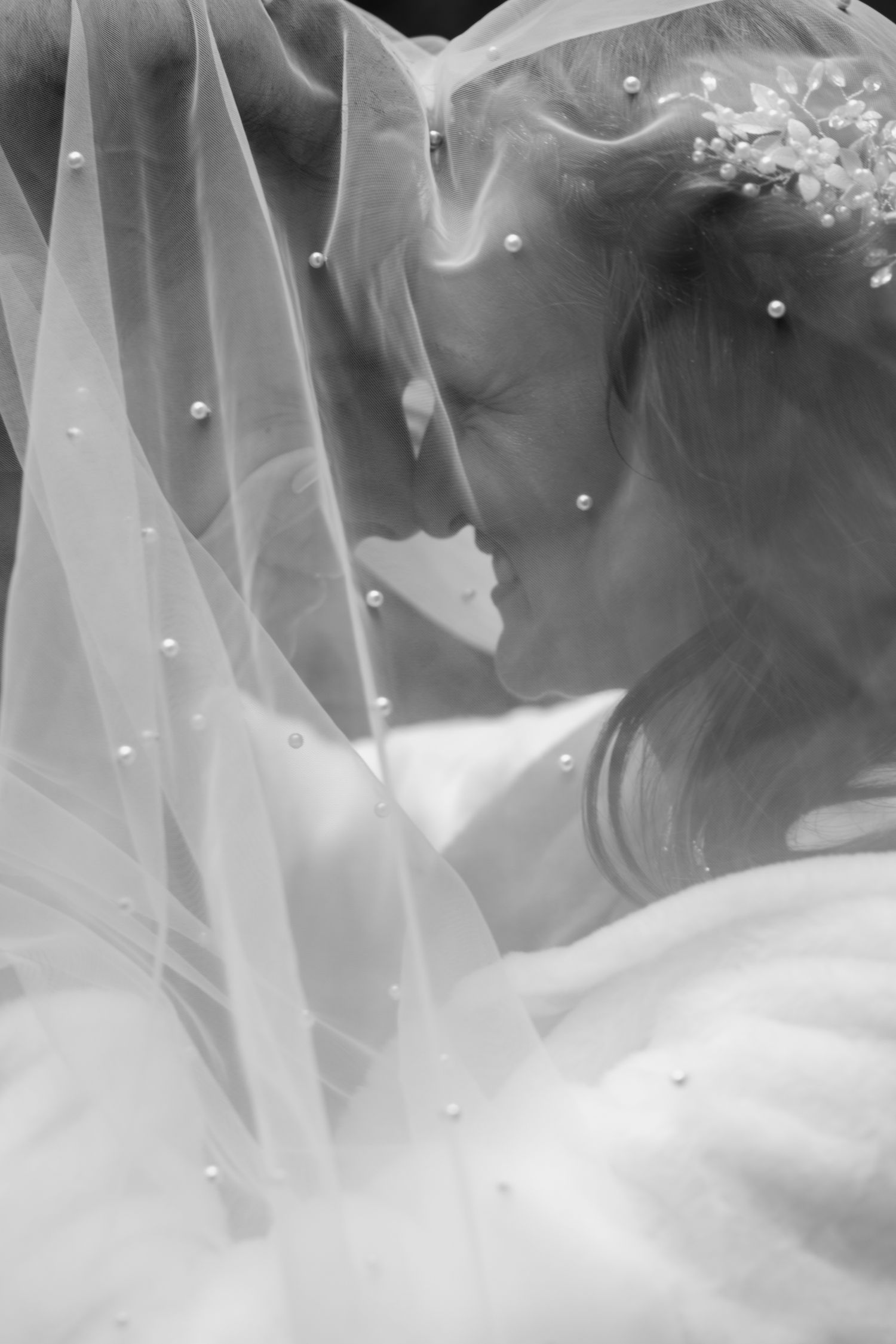 A romantic black and white wedding photo showing a couple embracing under a delicate veil with pearl details.
