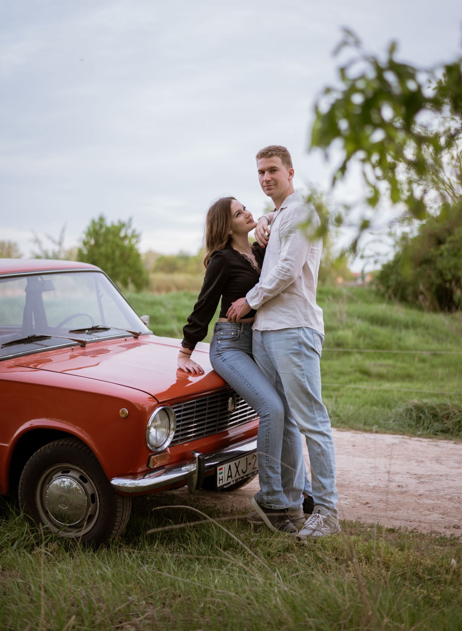 A romantic couple poses playfully by a vintage red car in a rural setting.