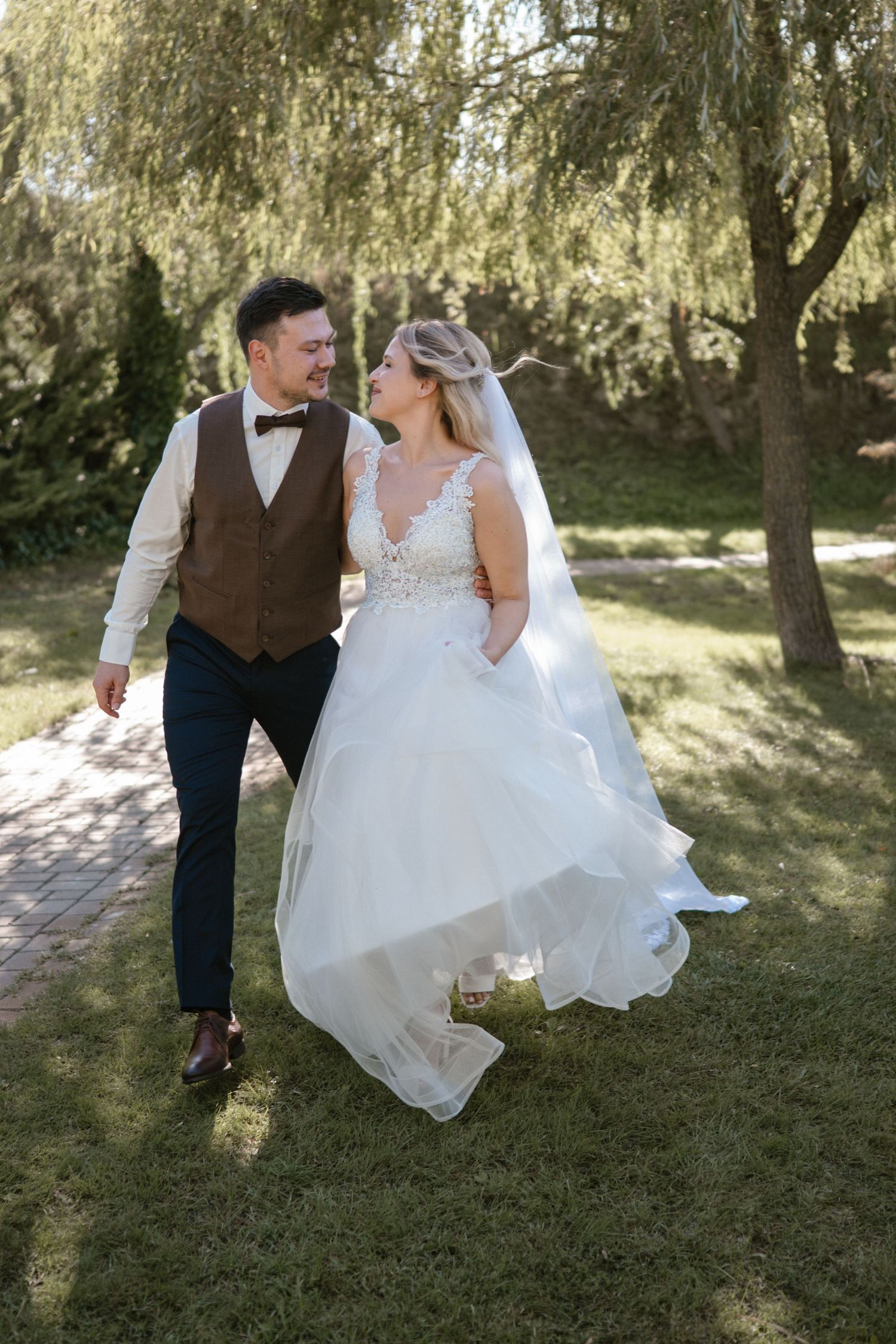 Couple walking together through a garden setting with willow trees, the bride's white dress flowing in the sunlight.