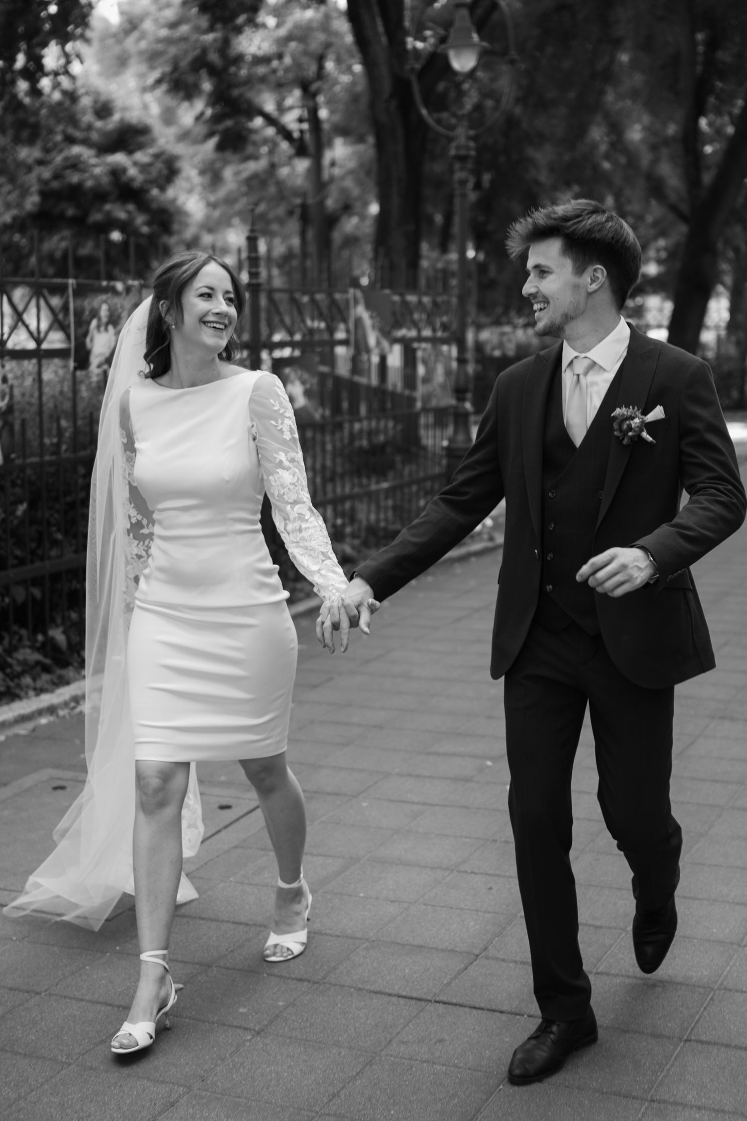 A bride and groom laugh while walking together on a tree-lined path.