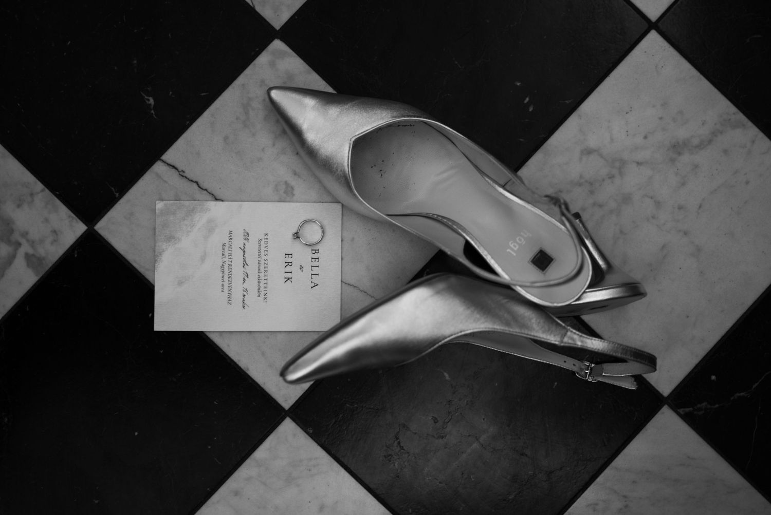 Black and white photo of silver pointed-toe heels beside an invitation card on a checkered marble floor.