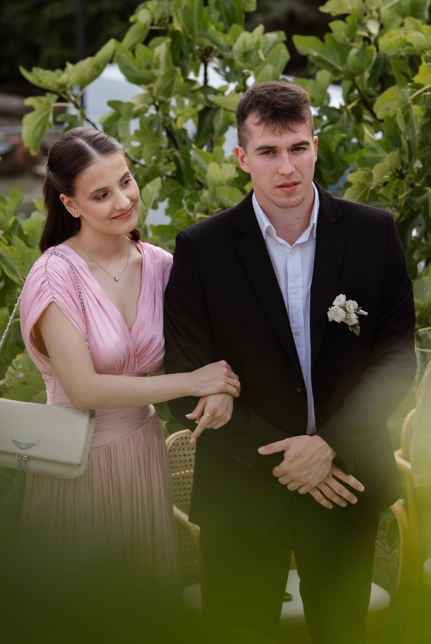 Young couple at a wedding, woman in pink dress and man in black suit with white boutonniere, posing outdoors.