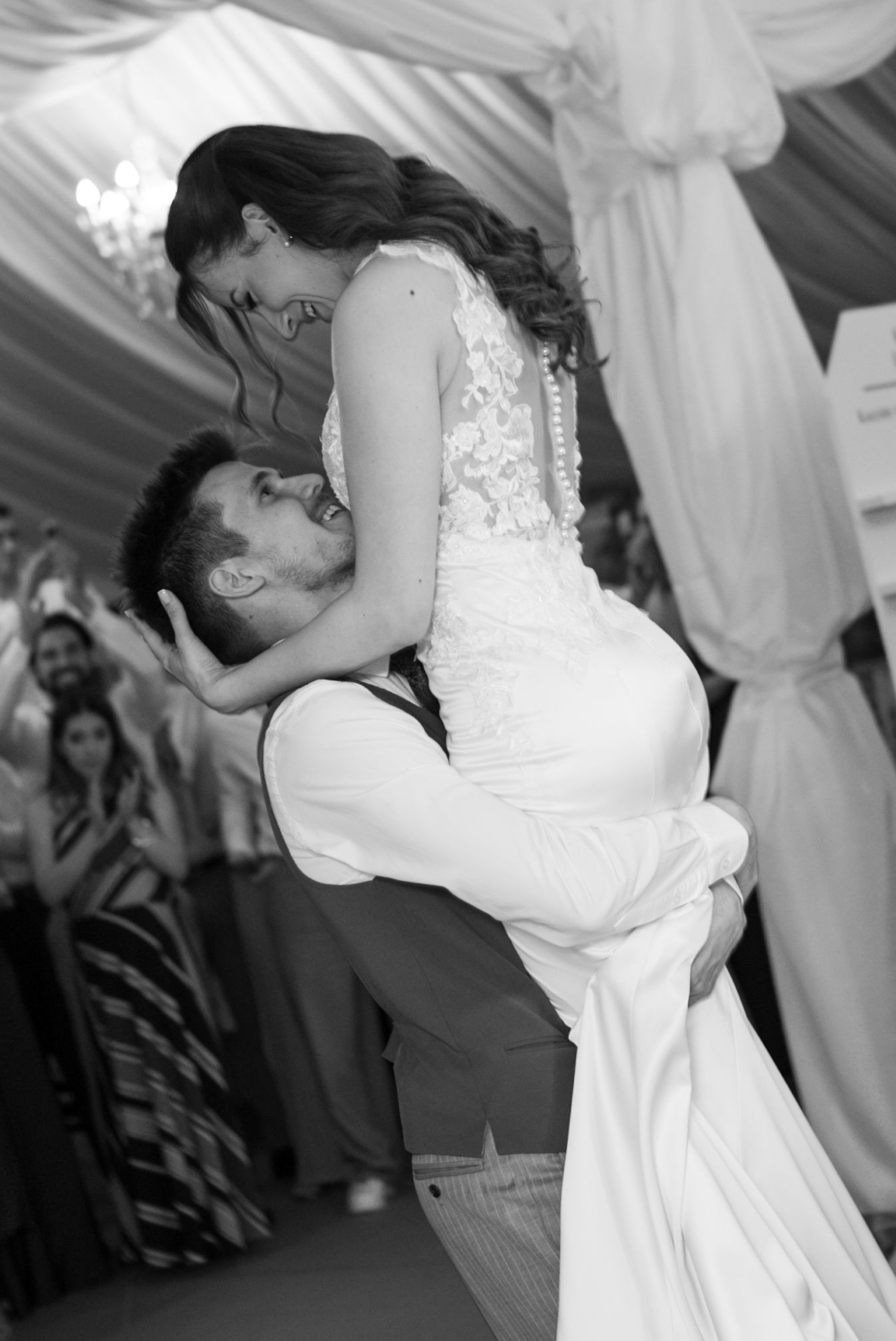 Groom lifting bride during first wedding dance in black and white photo with guests celebrating in background.