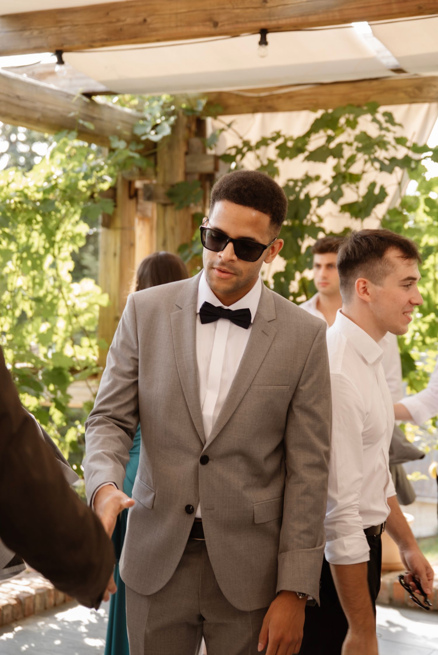 Stylish wedding guest in gray suit and black bow tie wearing sunglasses at an outdoor summer wedding.