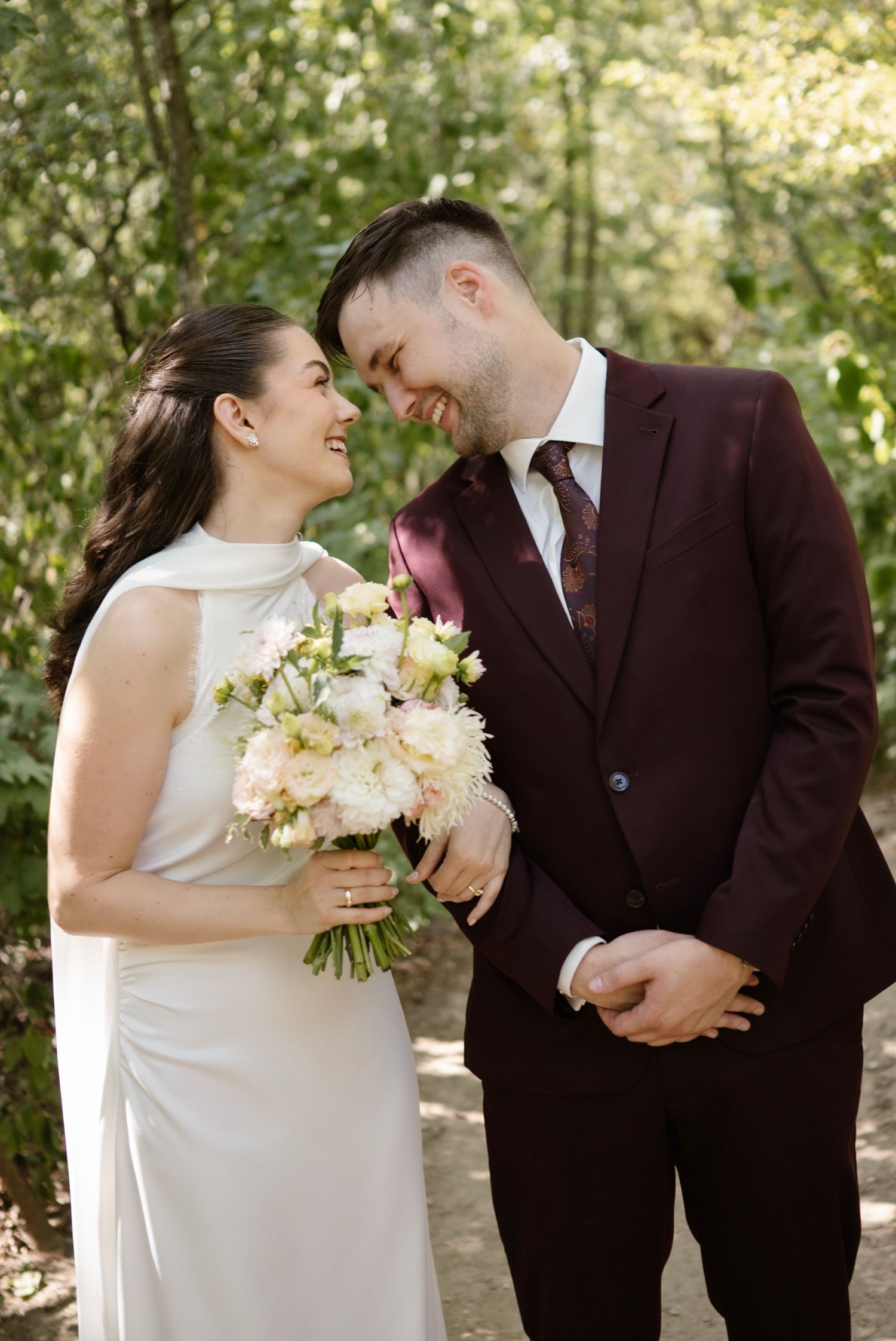 Smiling bride in white halter gown holds bouquet as groom in burgundy suit gazes at her outdoors.