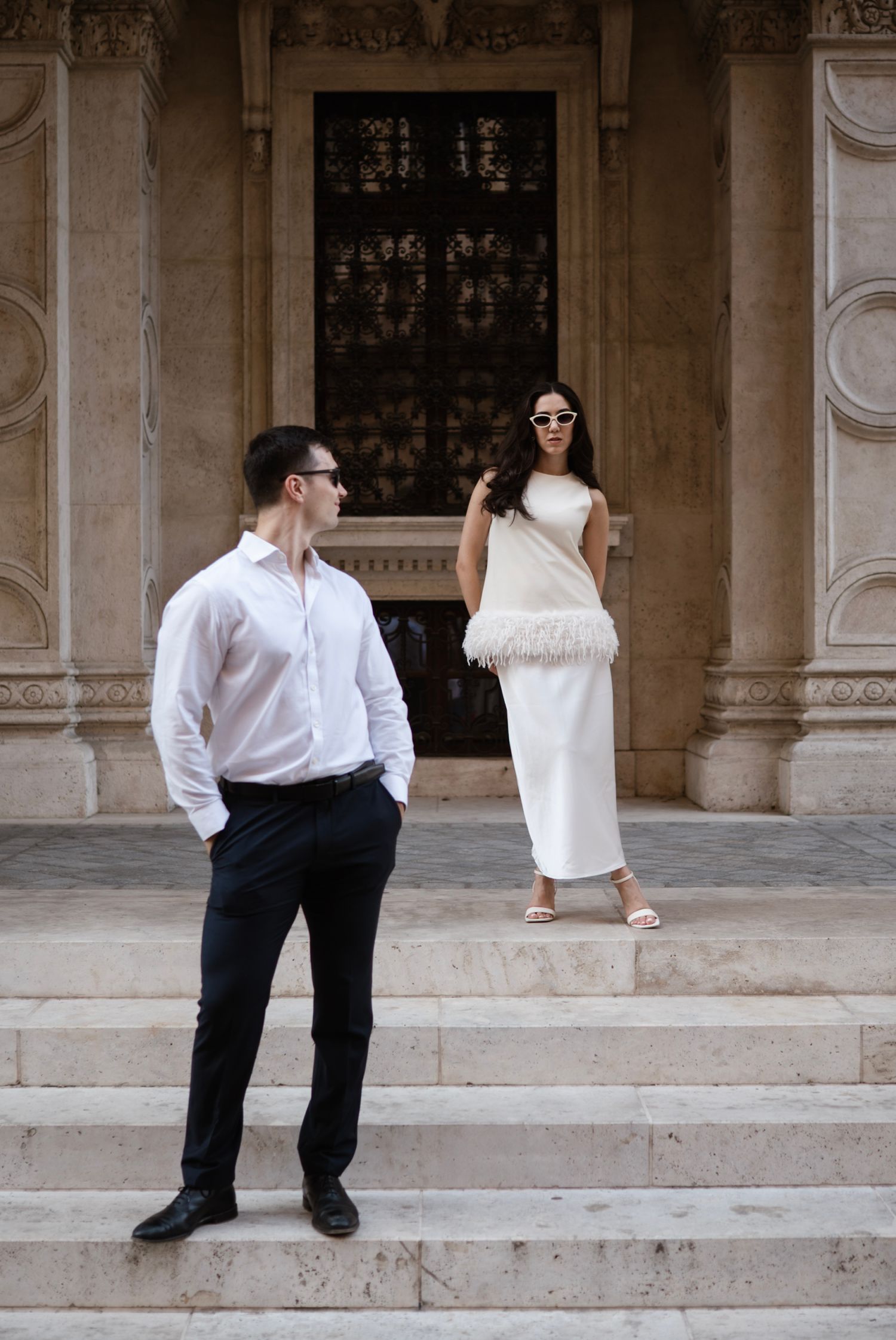 Elegant couple posing on marble steps, man in white shirt and black trousers, woman in white feather-trim dress with sunglasses.