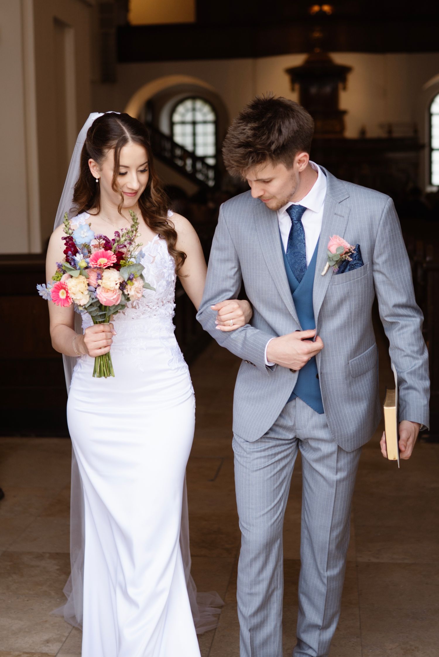 Bride in white gown holding colorful bouquet walks arm-in-arm with groom in gray suit inside elegant venue.