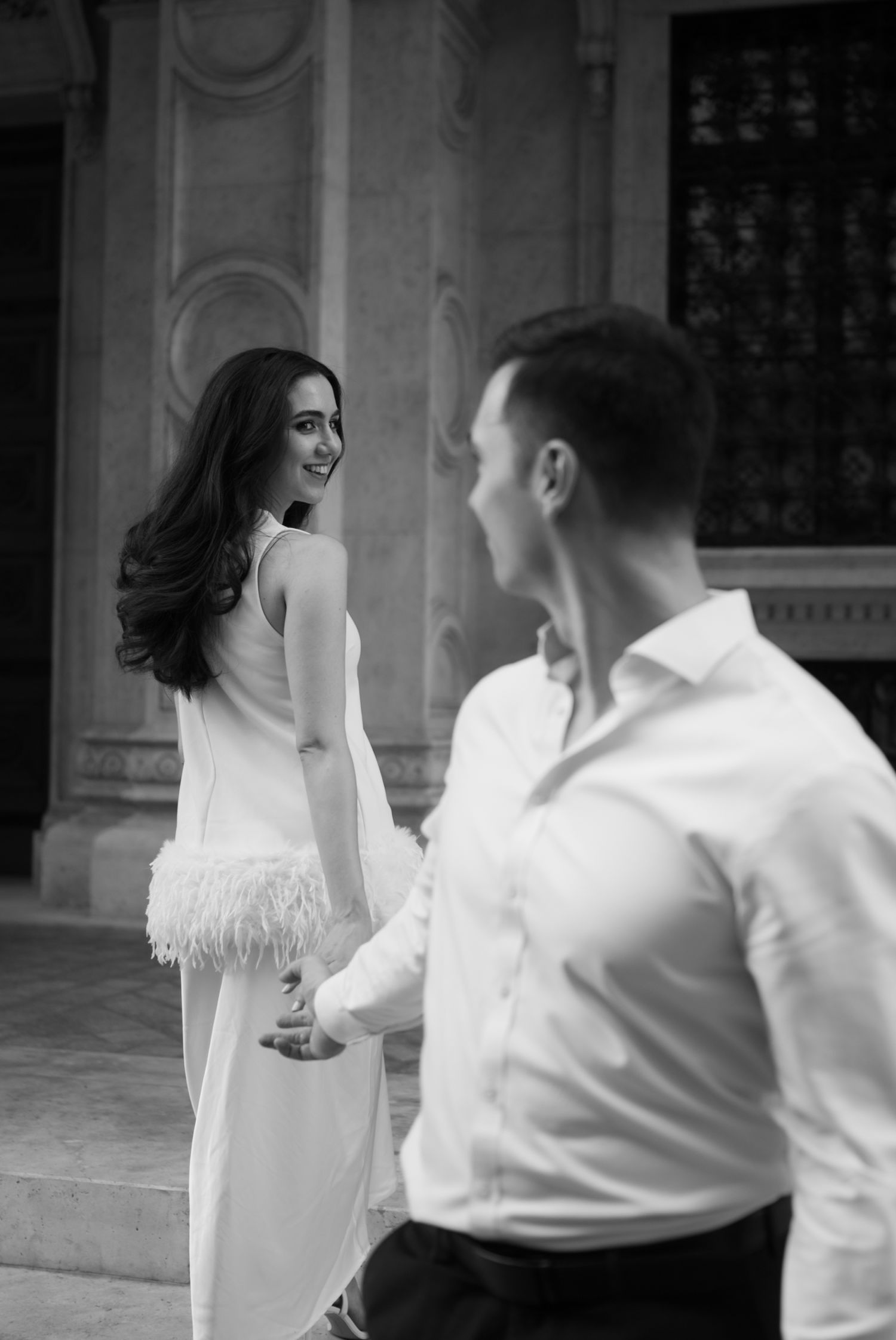 Black and white photo of a smiling woman in a white feathered dress holding hands with a man in a white shirt.