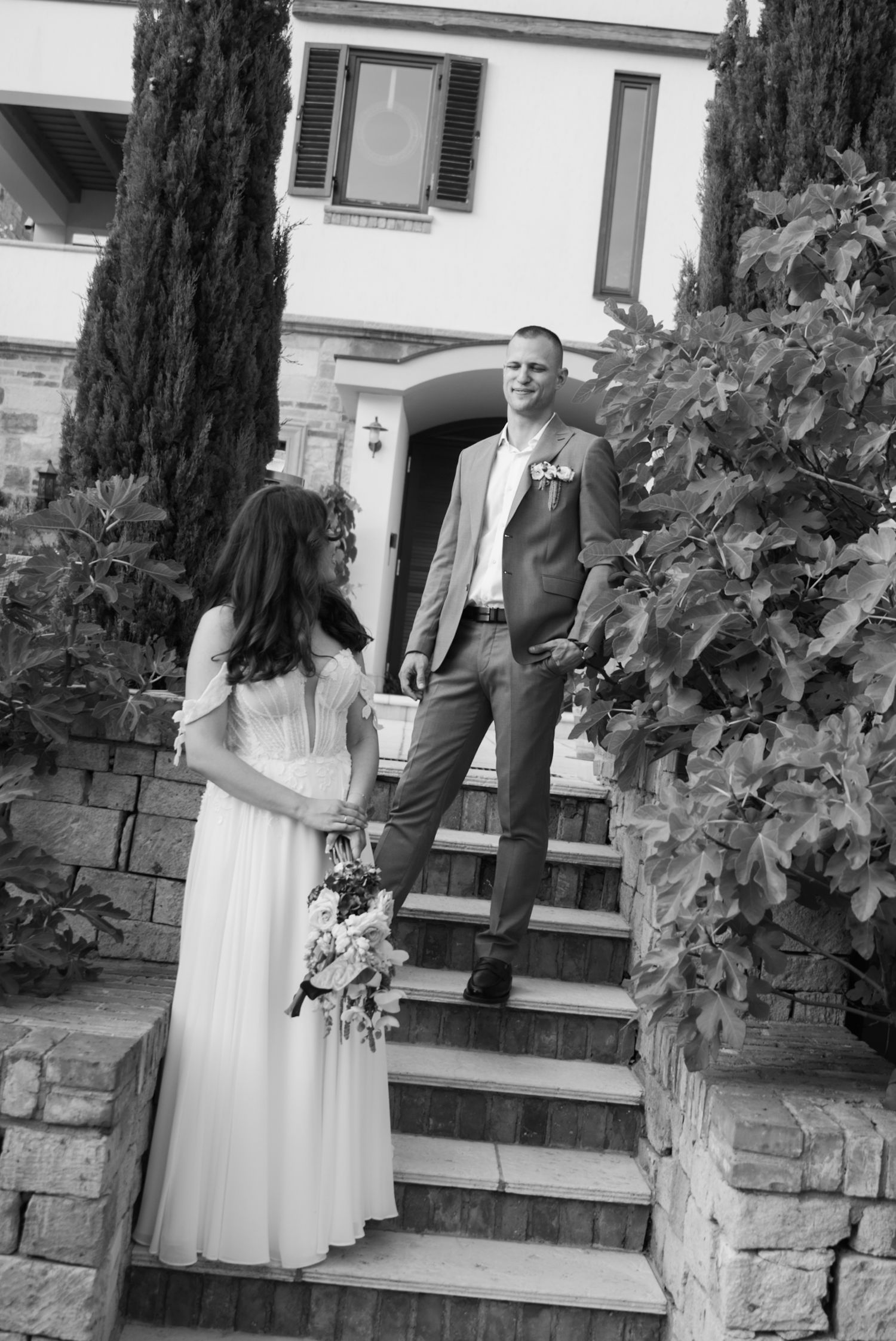 Black and white photo of a bride holding bouquet and groom in suit on stone steps outside a villa.