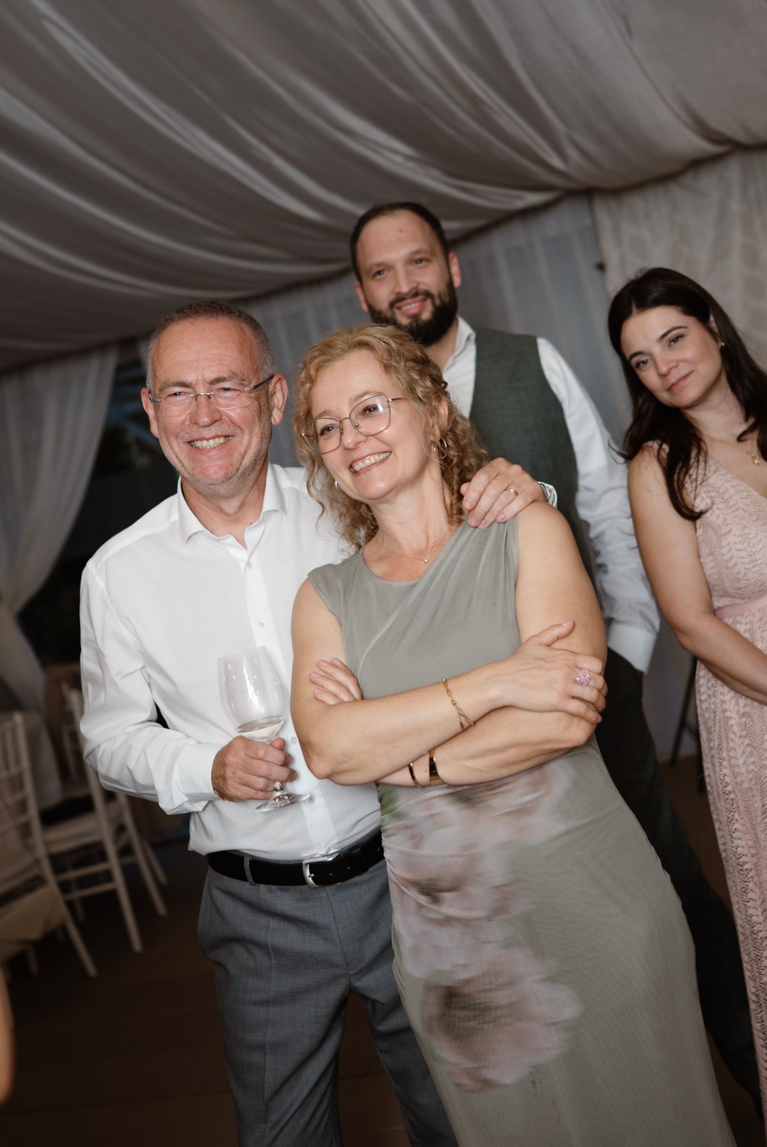 Four guests smiling at a wedding reception under a draped tent, one man holding a wine glass.