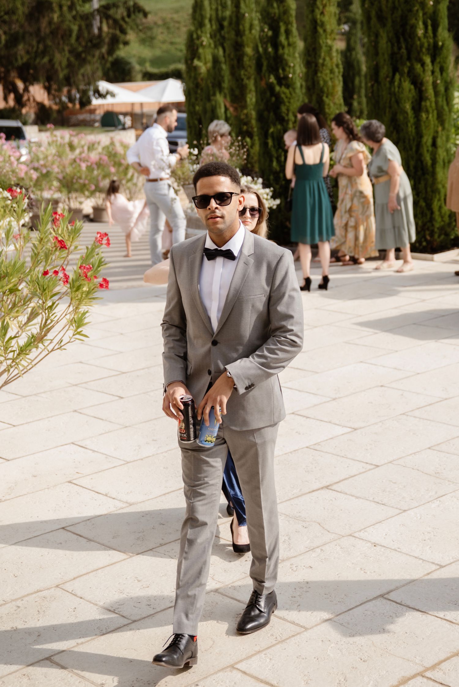 Dapper groomsman in gray suit and black bow tie with sunglasses holding wedding programs outdoors.