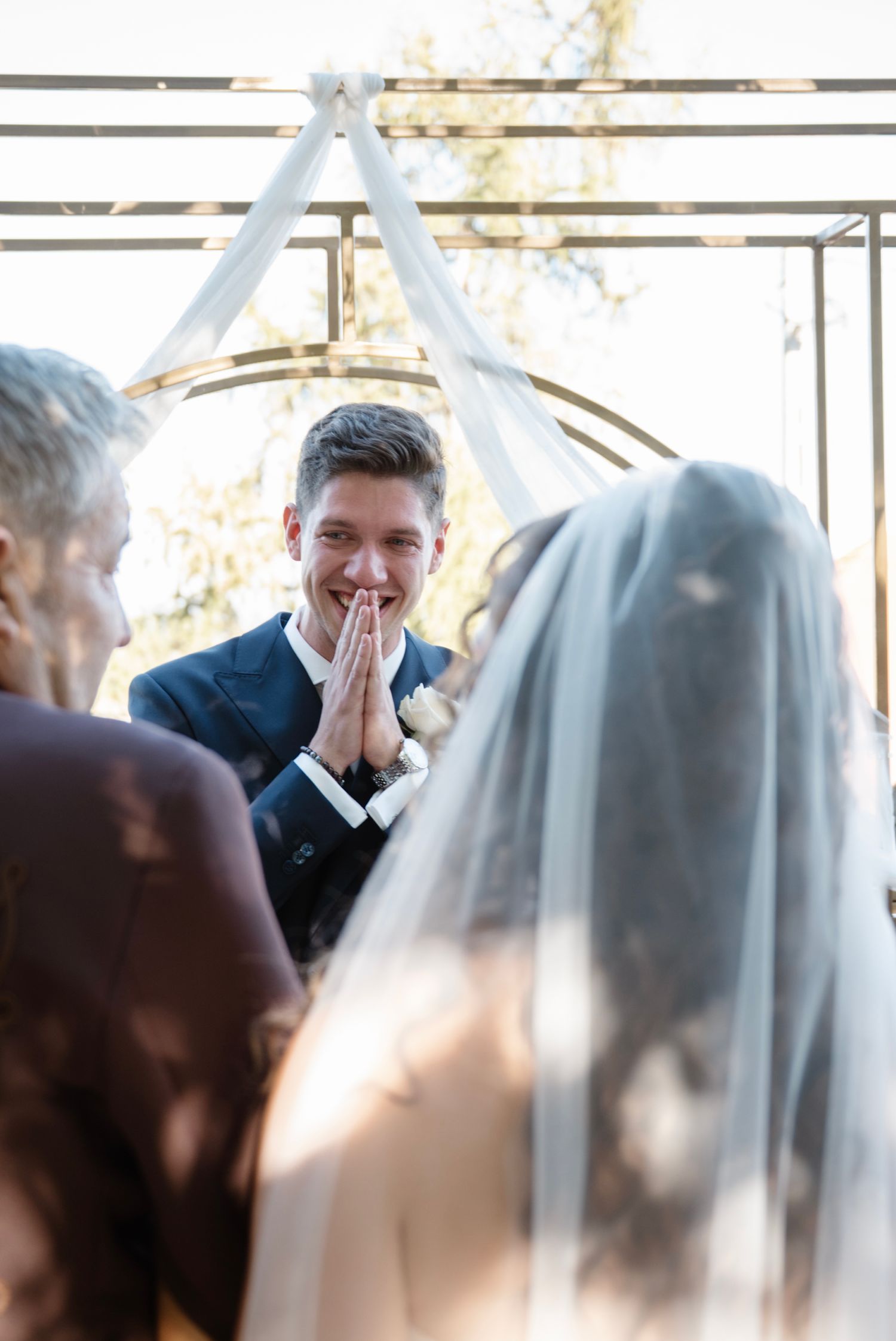 Groom clasps hands with emotional smile as bride approaches altar at outdoor wedding ceremony.
