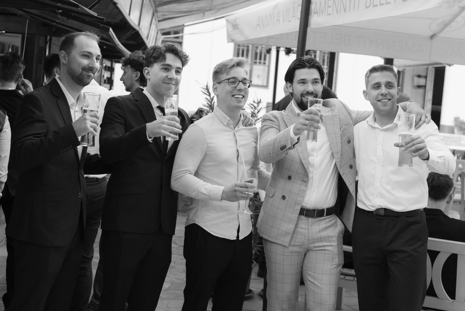 Group of five men in formal attire smiling and raising glasses in a celebratory toast at wedding reception.