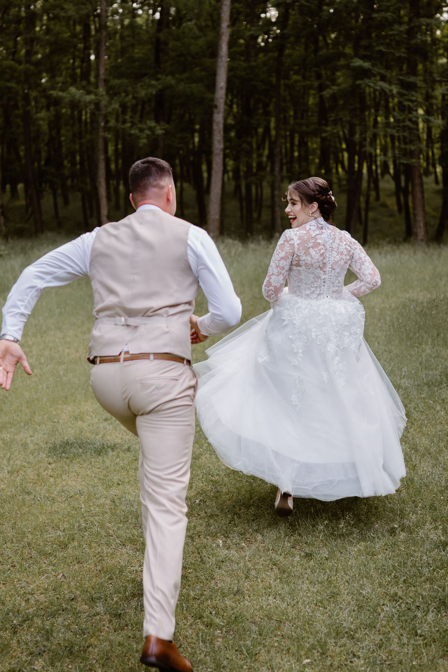 A couple in wedding attire playfully runs through a grassy field surrounded by trees.