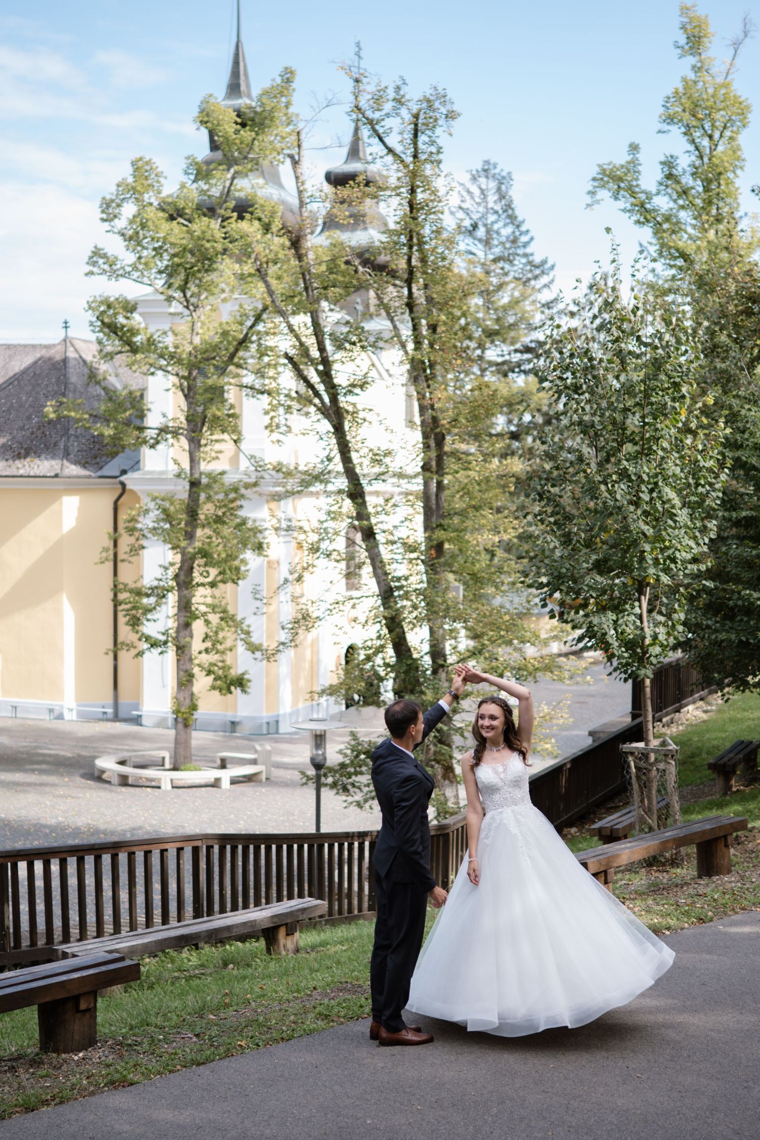 A couple dancing together outdoors in front of trees and a fence, the bride wearing a flowing white ballgown.