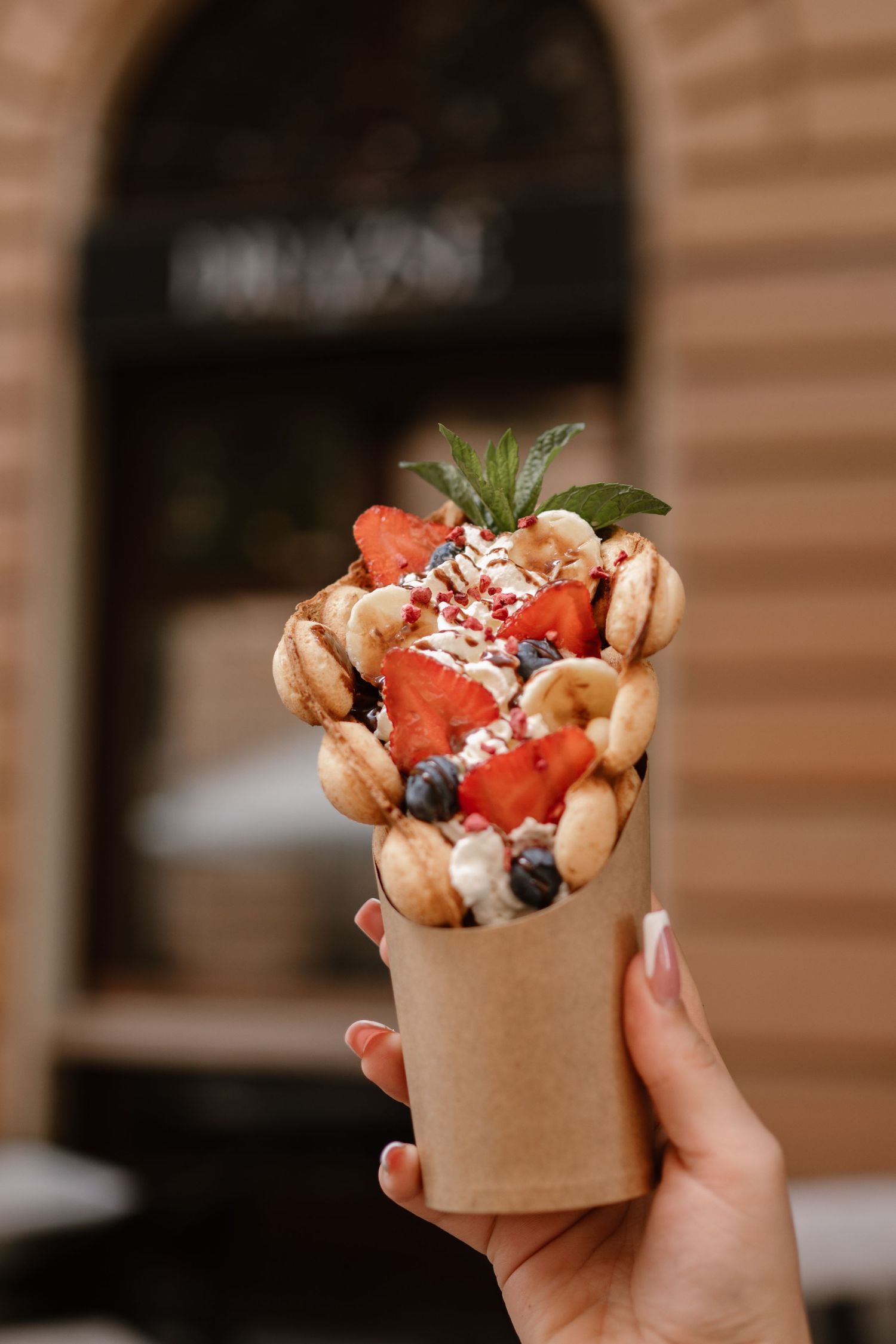 Hand holding bubble waffle cone filled with fresh strawberries, blueberries and whipped cream against wooden background.
