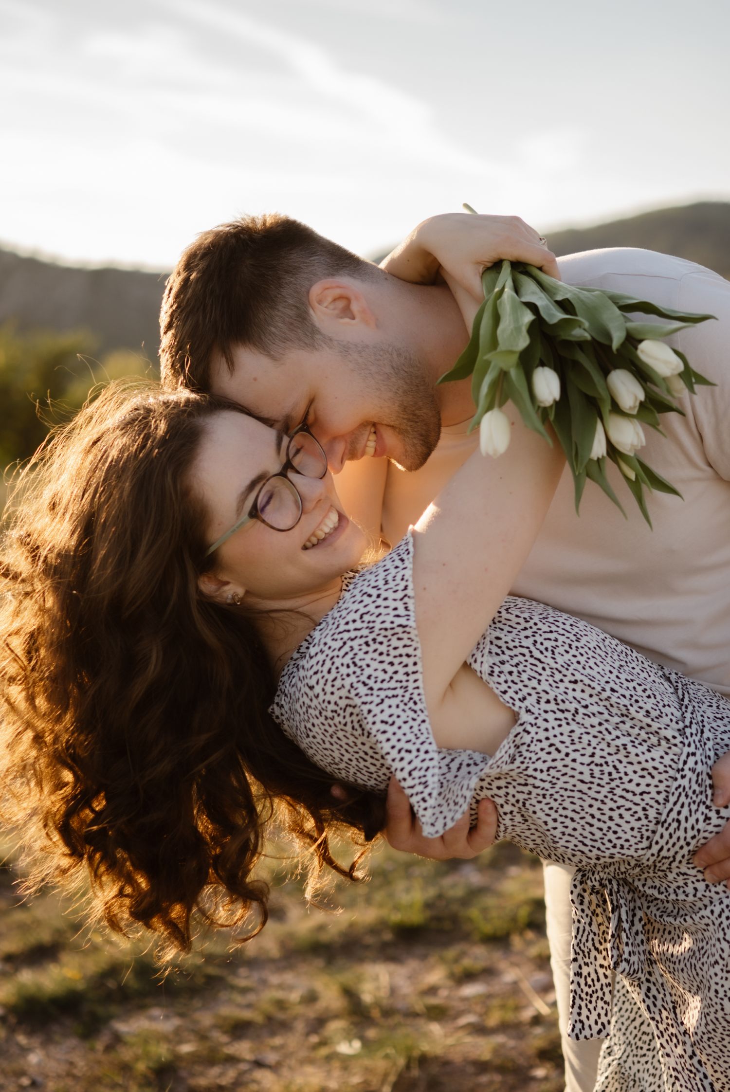 Couple sharing romantic moment at sunset, man dipping woman holding white tulips in scenic outdoor setting.