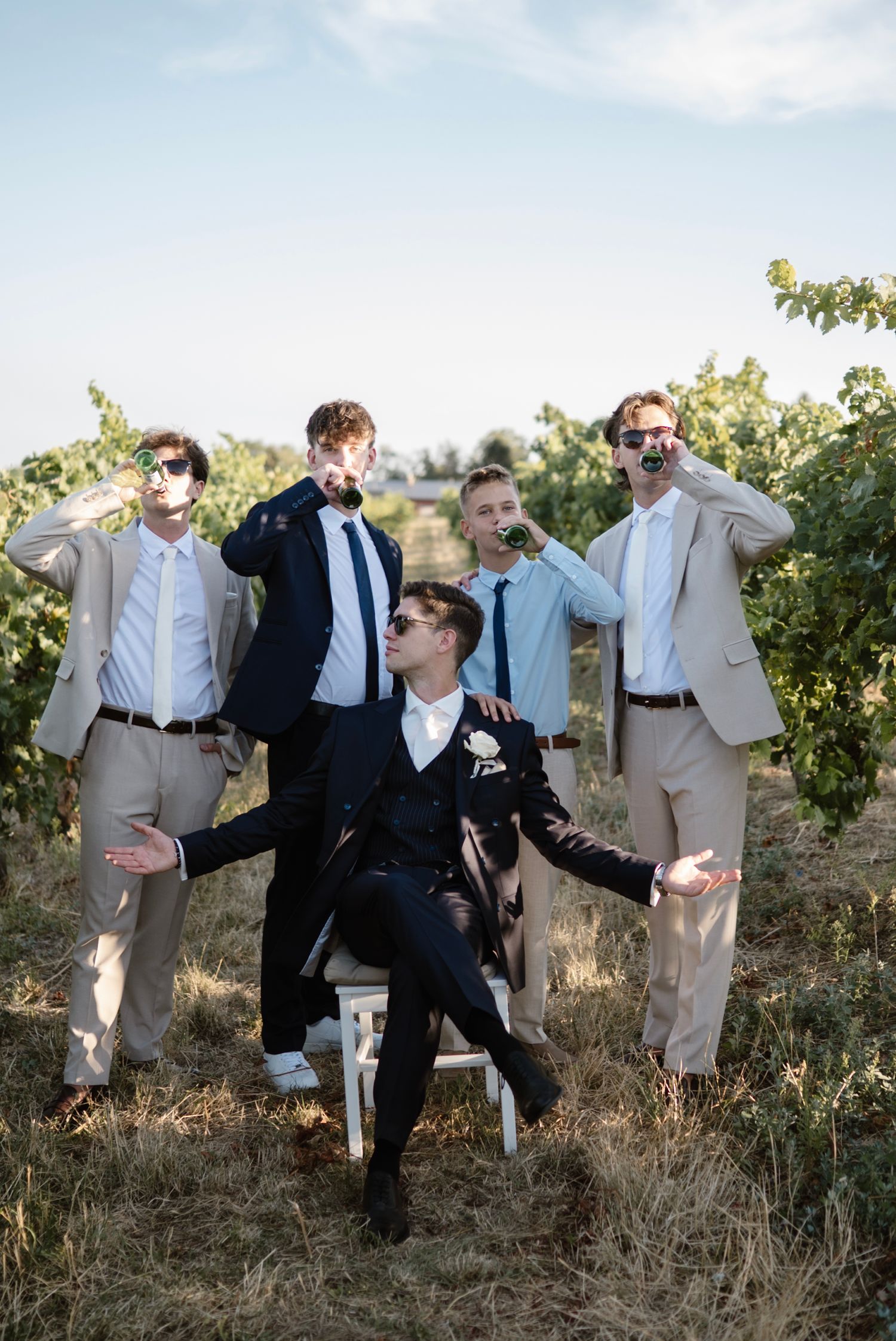 Groom in black suit seated with groomsmen in tan suits drinking wine in a vineyard on a sunny day.