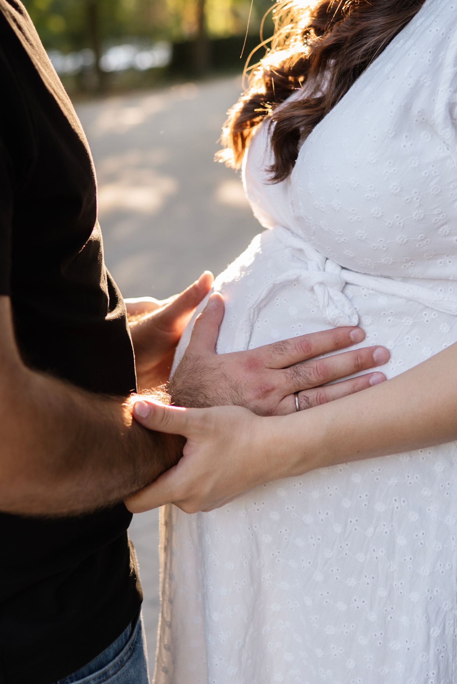 Couple tenderly touching pregnant belly outdoors in sunlight, woman in white dress with partner's hand over hers.