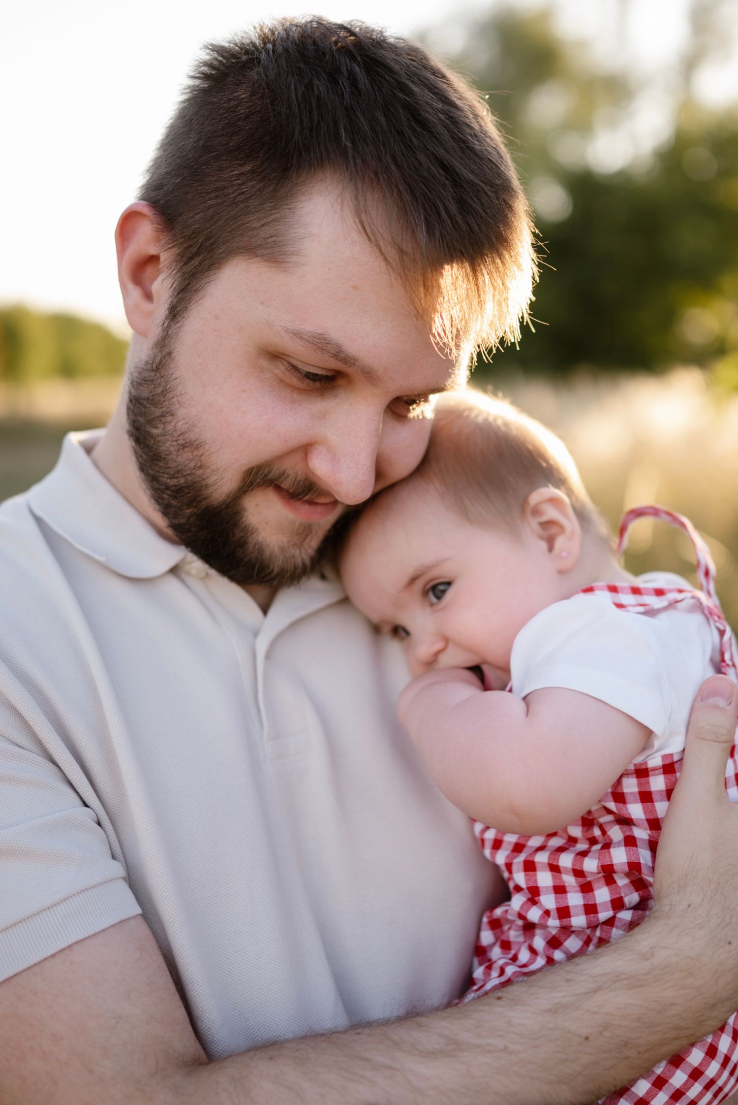 A bearded father tenderly holds his baby in a red gingham outfit outdoors in golden sunlight.