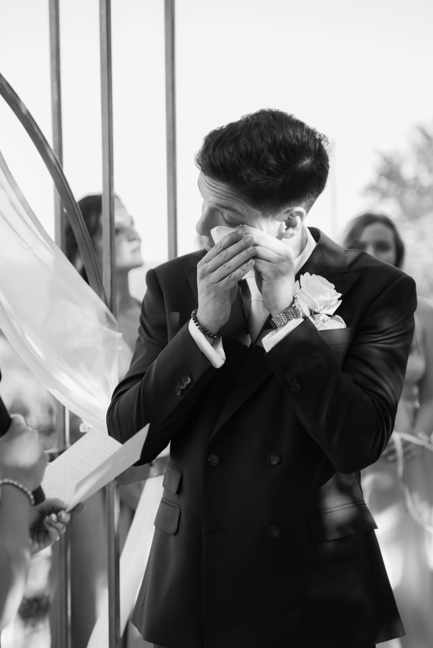 Tearful groom in black suit wiping eyes with boutonniere rose at outdoor wedding ceremony.