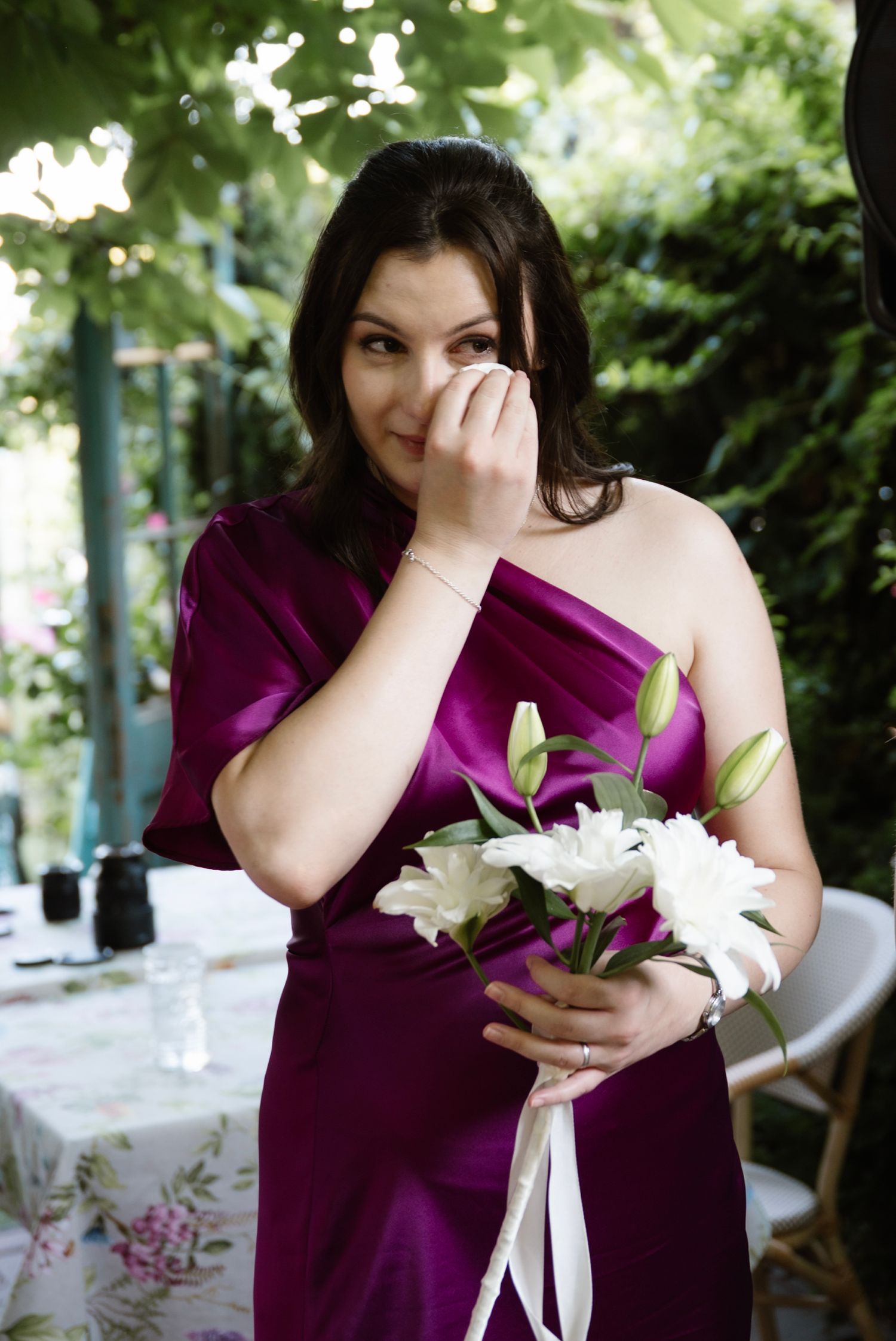 Tearful bridesmaid in purple satin dress holding white lily bouquet at wedding reception.