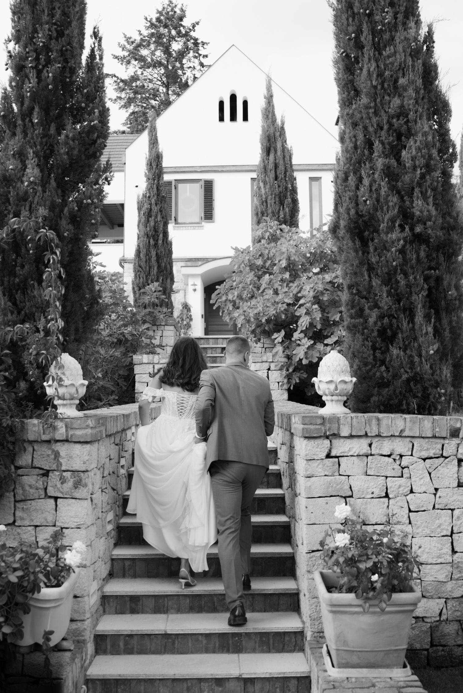 Black and white photo of a couple walking up stone steps toward an elegant villa surrounded by tall cypress trees.
