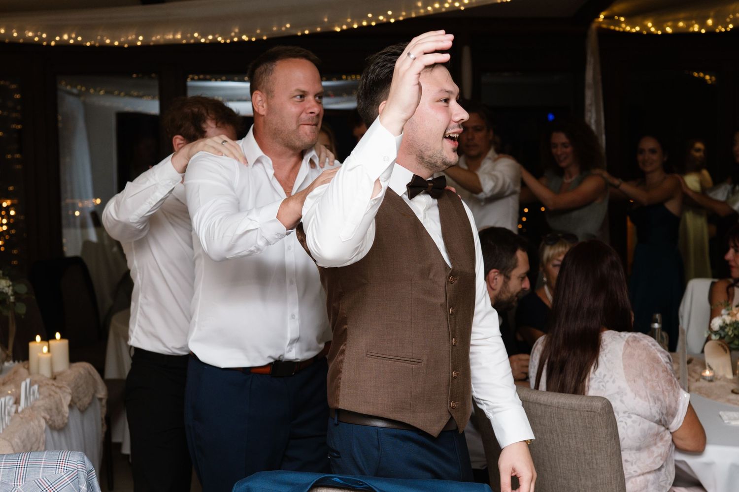Wedding guests dancing and celebrating under warm string lights at an indoor reception.