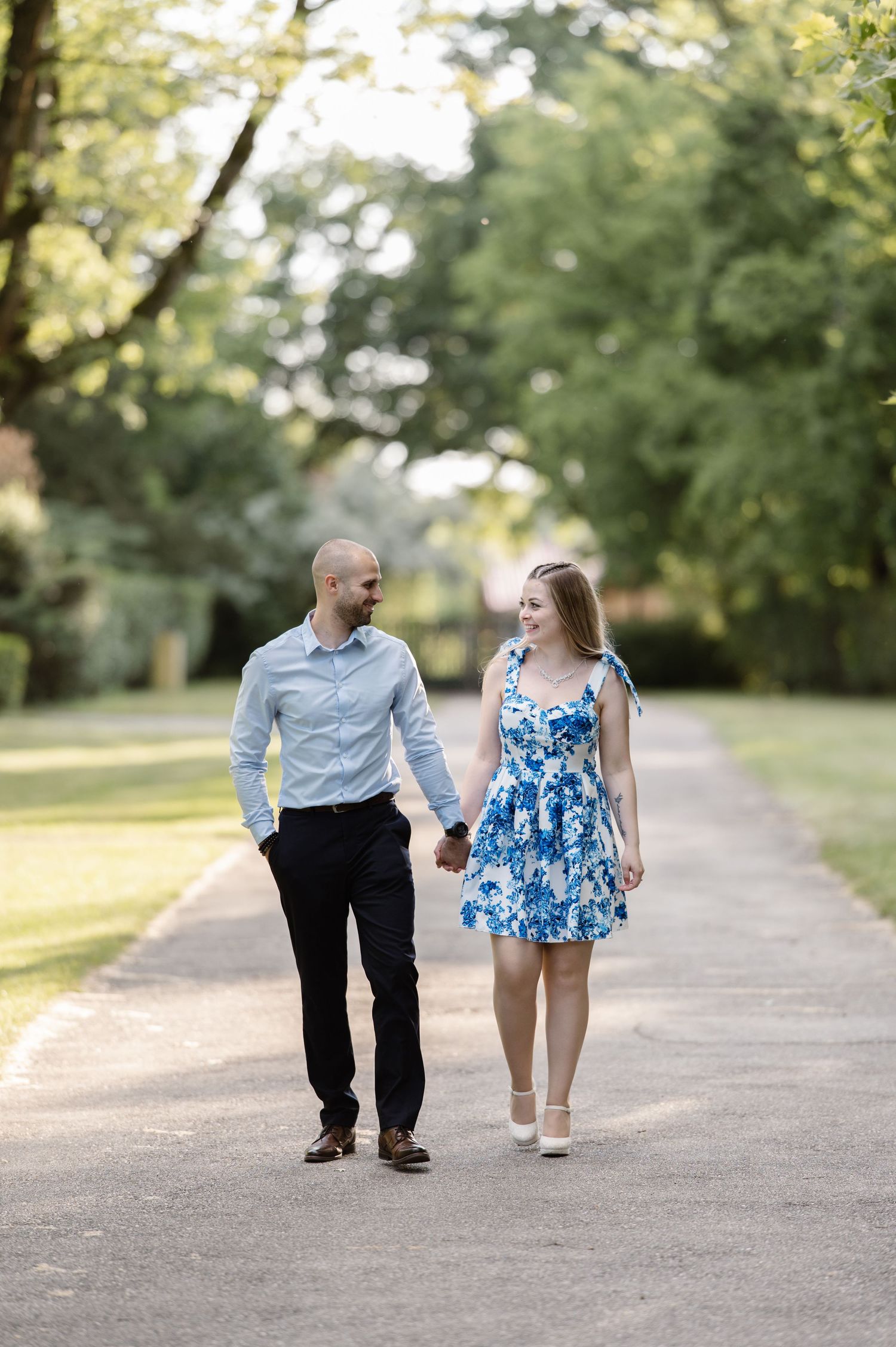 Couple walks hand in hand down a tree-lined path during summer photo session.
